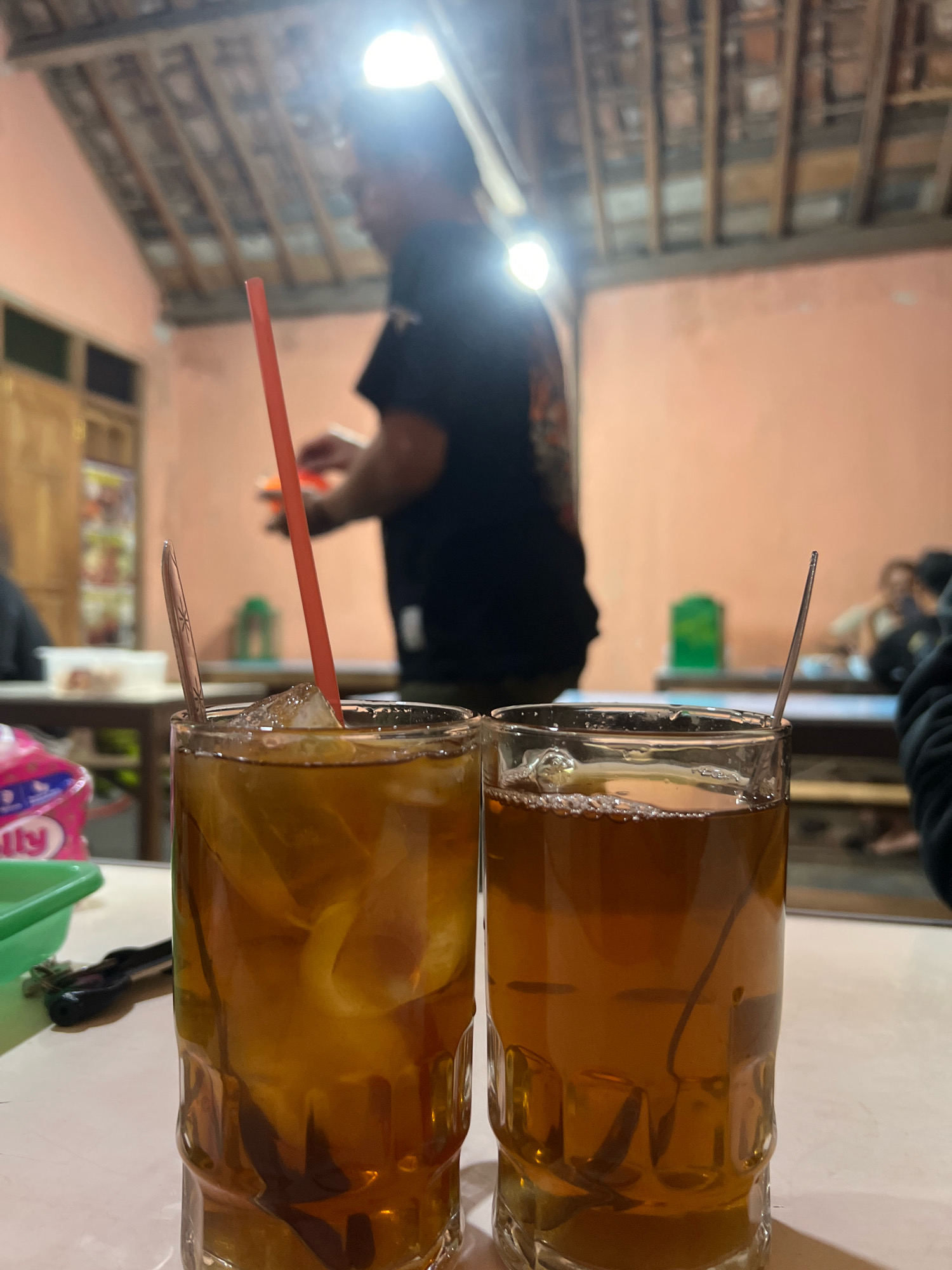 In the foreground, two glasses of iced tea sit on a white table. The glass on the left contains ice, lemon slices, and a red straw. The glass on the right has a spoon and a tea bag. In the background, a blurred figure stands near an open doorway, suggesting an indoor setting with some form of rustic architecture, possibly a traditional Javanese house or a warung (food stall). The lighting appears to be artificial, with a bright light source illuminating the ceiling beams. The walls are painted a light orange. Based on the overall aesthetic, this scene is likely located in Sleman, Indonesia, a region known for its traditional culture and eateries.
