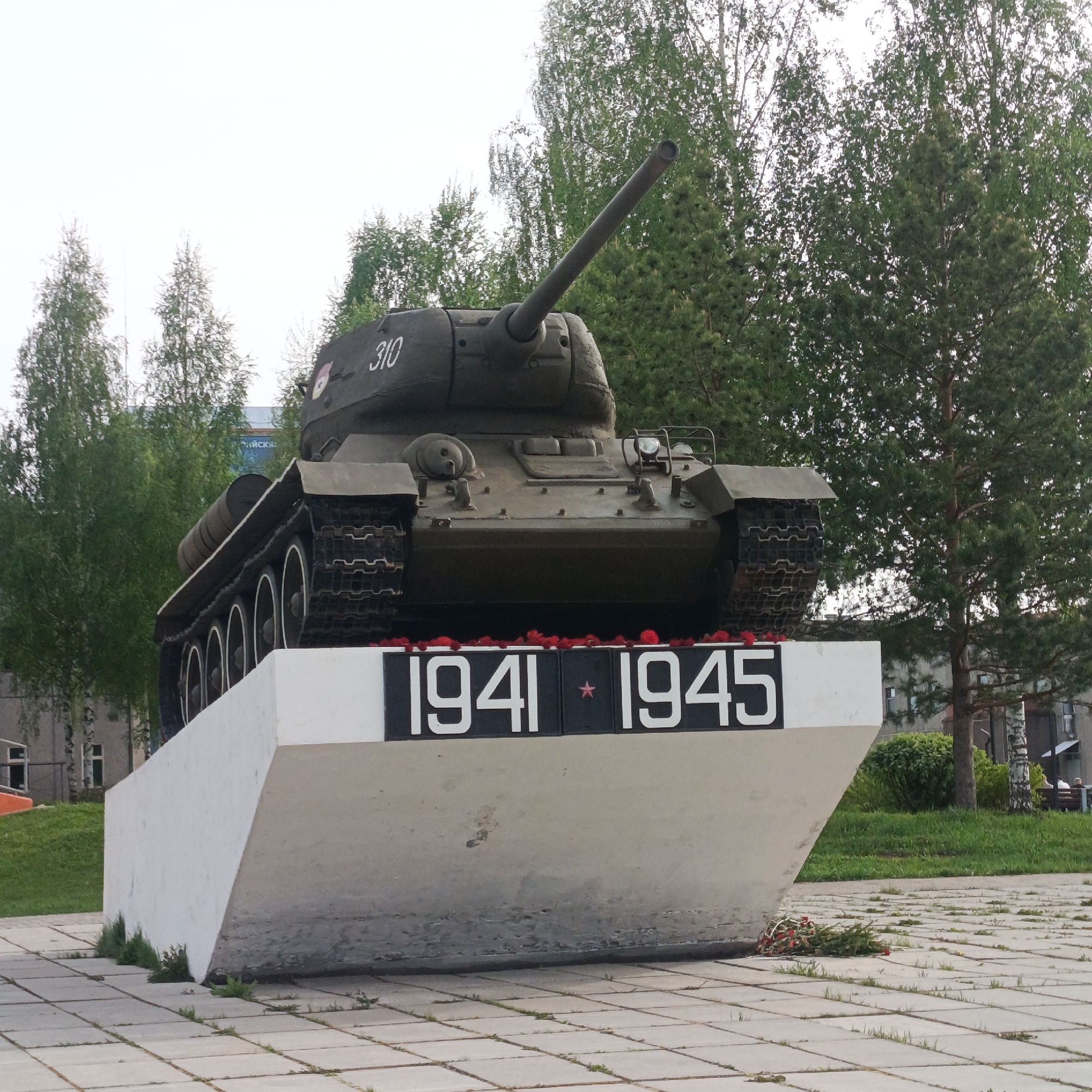 A T-34 tank is mounted on a white concrete plinth in a city park in Марковский, Russia. The plinth has "1941 * 1945" painted in white on black rectangles. Red flowers are placed on the plinth and on the ground. The tank is painted green and has the number "310" on the turret. Trees are in the background, and a building is visible behind the trees. The ground is paved with light gray tiles.