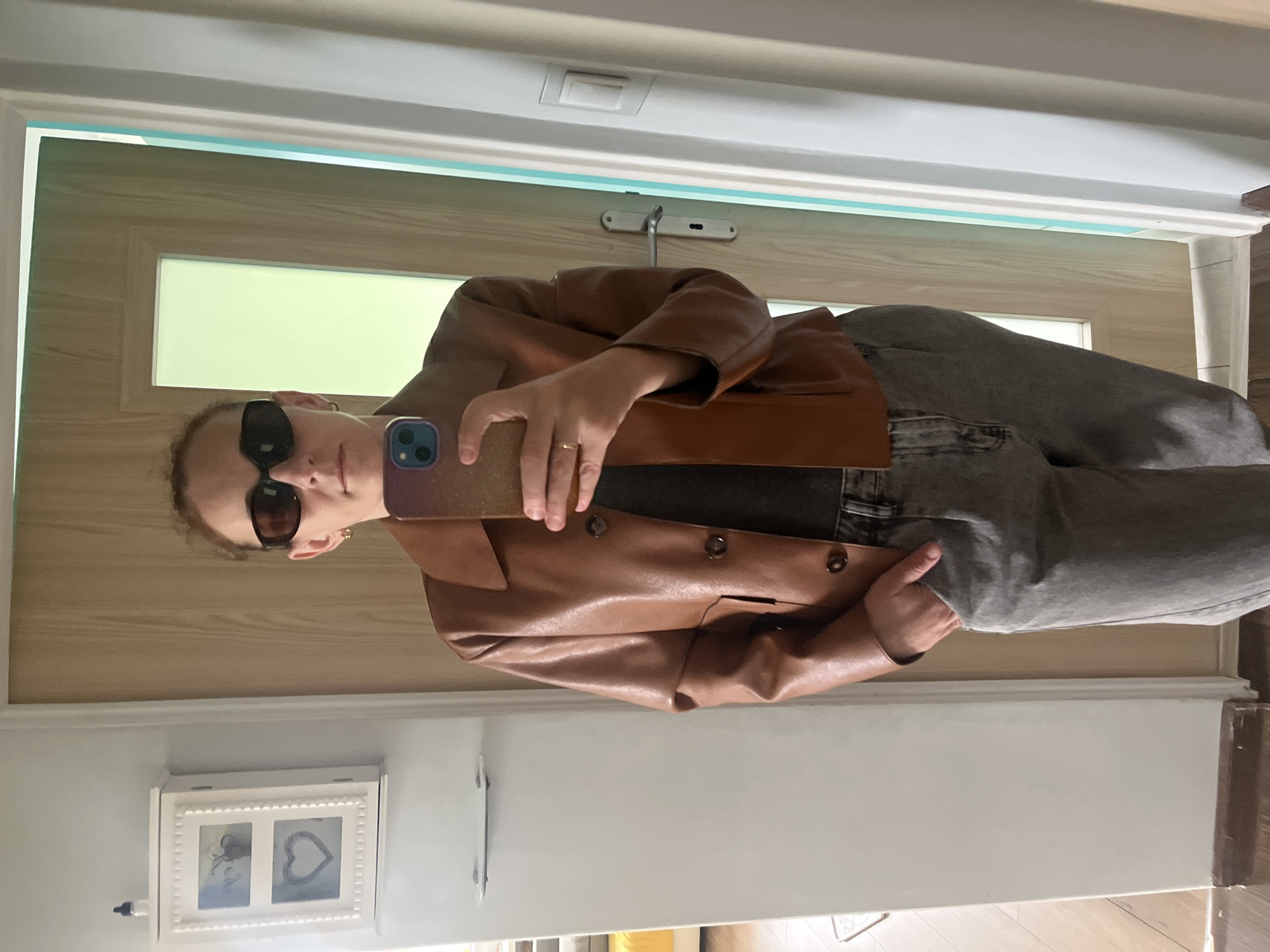 A selfie taken in a bathroom of a woman wearing sunglasses, a tan leather jacket, and gray pants. The background consists of a door, wall, picture frame and the bathroom floor. Location: Unknown, Unknown.