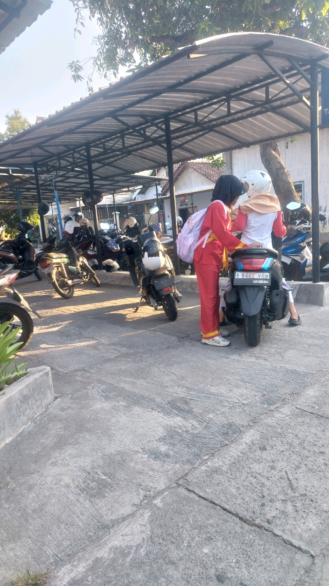 In Ronowijayan, Indonesia, a low-angle, outdoor, medium shot captures a paved area with several parked motorcycles beneath a corrugated metal canopy supported by metal poles. Two young females, appearing to be students, are the primary subjects. One, wearing a red outfit and a headscarf, stands beside a dark motorcycle, holding a pink backpack with a white strap. The other female, seated on the motorcycle, wears a white top, a tan head covering, and a white helmet. The motorcycle has a license plate that reads "B 6442 VBW" with a date stamp below. Other motorcycles are visible in the background. The environment suggests a parking lot or a schoolyard, indicated by the parked vehicles and the presence of young individuals in uniform-like attire. The lighting suggests it is late afternoon.