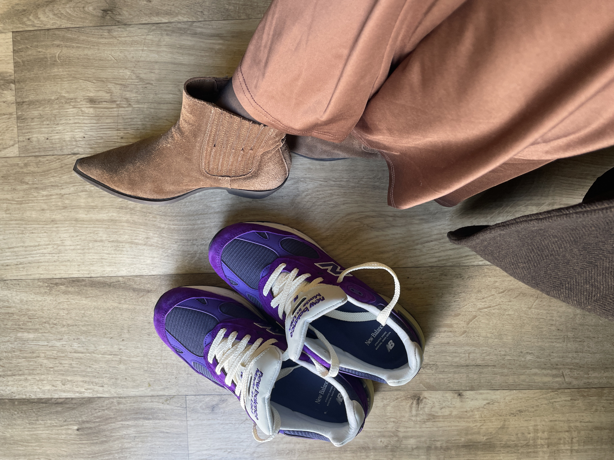A high-angle shot shows a pair of brown suede cowboy boots worn by someone in copper-colored pants, next to a pair of purple and gray New Balance sneakers, placed on a light wood floor. A brown hat lies to the side.