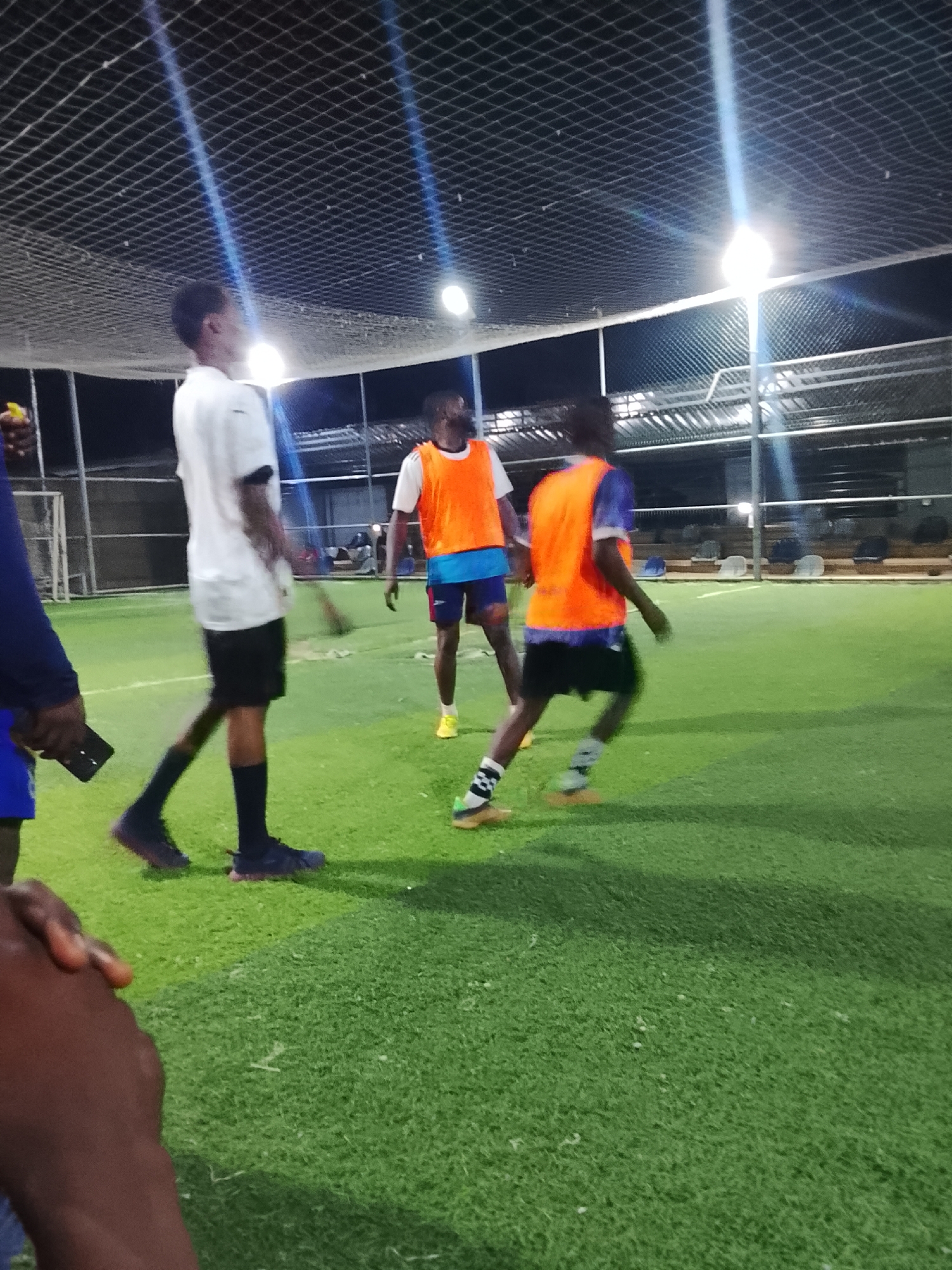 The image shows a group of young men playing soccer on an artificial turf field at night in what appears to be a caged or enclosed area. Overhead netting is visible, along with several bright lights illuminating the field. One man wears a white shirt and black shorts, and another wears an orange practice vest over a blue shirt and shorts. A third man stands in the background wearing an orange vest. Several other people are partially visible in the foreground, with one holding a phone. Numerous chairs are visible in the background. While the image is provided, it is not possible to confirm that this is from Akwanga, Nigeria.