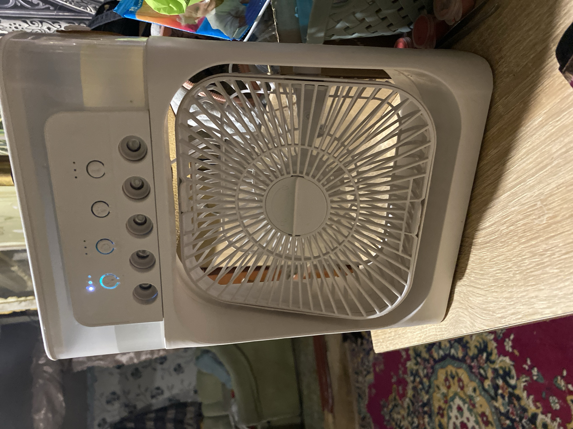 An indoor, eye-level, medium shot captures a white portable evaporative cooler with a fan on a wooden table. The cooler has a clear top compartment filled with water and a control panel with several buttons and indicator lights. The fan, with its white grille, is positioned in the lower front of the unit. The background shows a dimly lit room with furniture and a patterned rug. The scene is devoid of people and any overt action. The context of Nova Praha, Ukraine, is not visually apparent in the image itself.