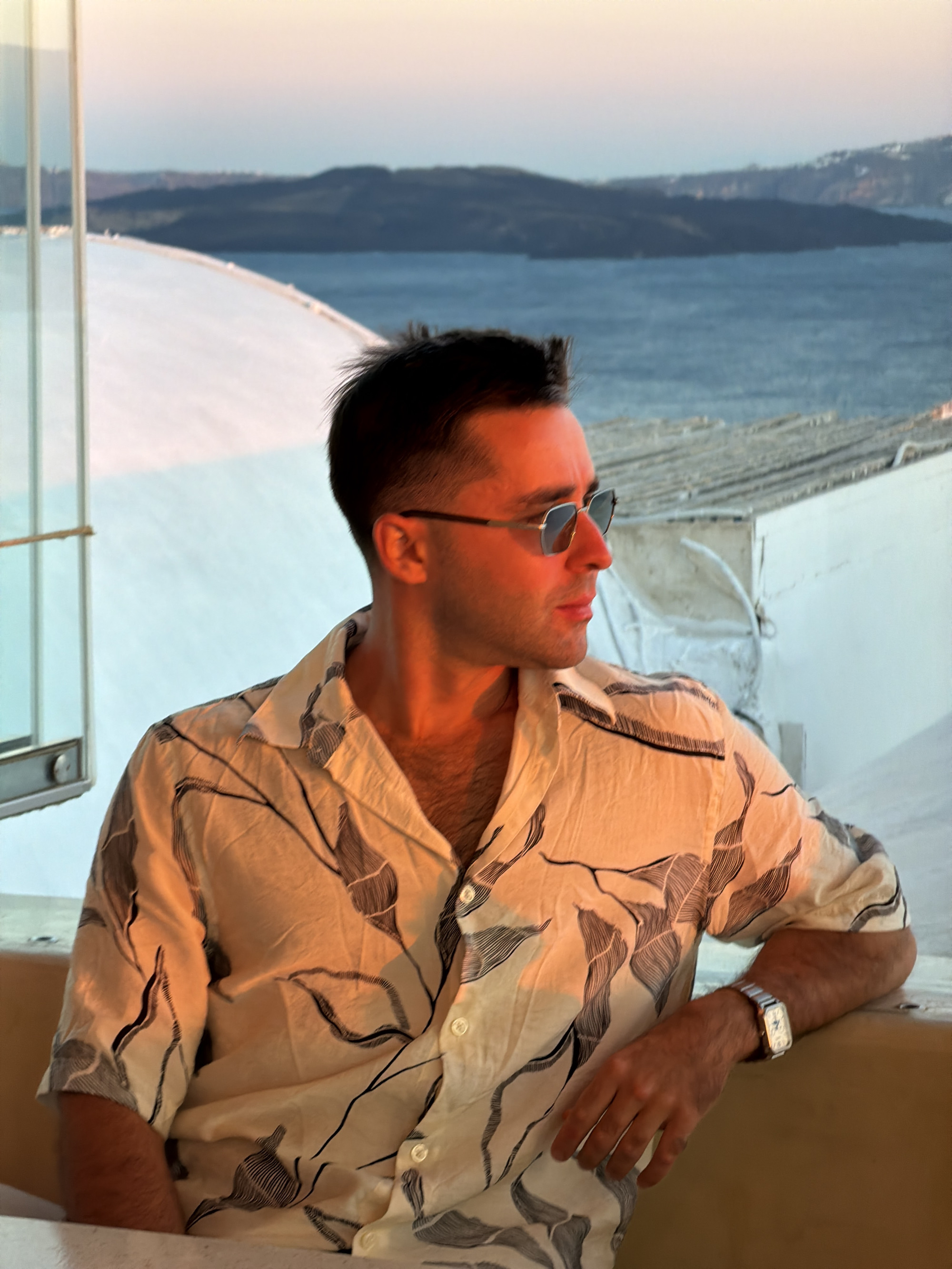 The image shows a man wearing a patterned button-up shirt and sunglasses, sitting outdoors with a view of the sea and hills in the background. The setting appears to be Ia, a town in Greece known for its white-washed buildings and stunning sunsets over the Aegean Sea. The man is looking off to the right, his arm resting on a railing. The lighting suggests it is either sunrise or sunset, casting a warm glow on his skin and shirt. The environment includes white structures, possibly buildings or terraces, and the blue sea with distant landmasses.