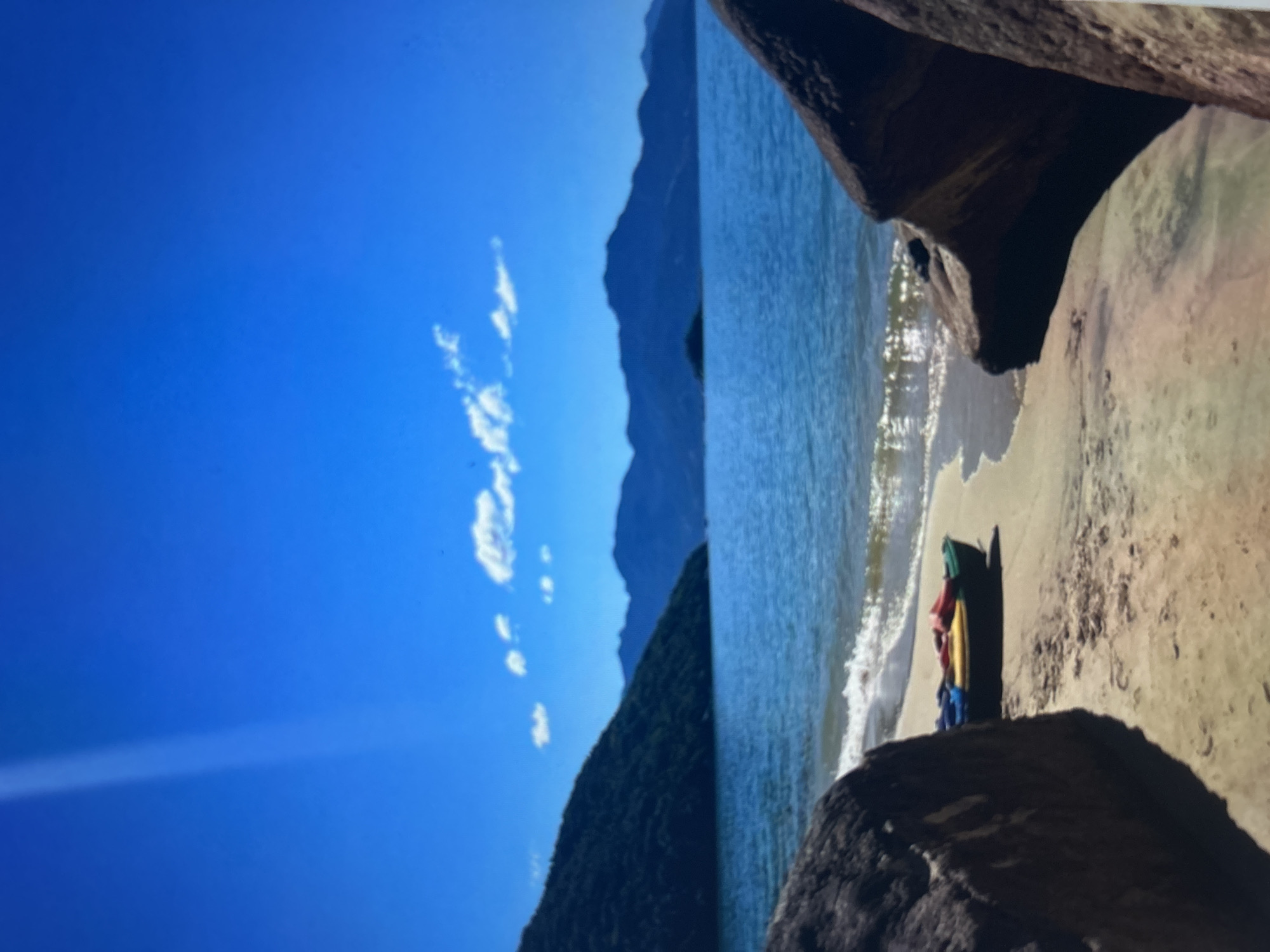 This is a high-angle, full shot of a serene beach scene on a clear, sunny day. In the foreground, two large, dark rocks frame the view. Between them, a sandy beach meets the calm, blue ocean. A small, colorful kayak, with red, yellow, and blue components, rests on the sand near the waterline. In the mid-ground, a lush green, tree-covered mountain slopes down to the sea on the left side of the image. The horizon is dominated by a range of blue mountains, fading into the distance under a vibrant, cloud-streaked blue sky. A faint streak of light, possibly from the sun, cuts across the upper left portion of the sky. The overall scene conveys a sense of tranquility and natural beauty.