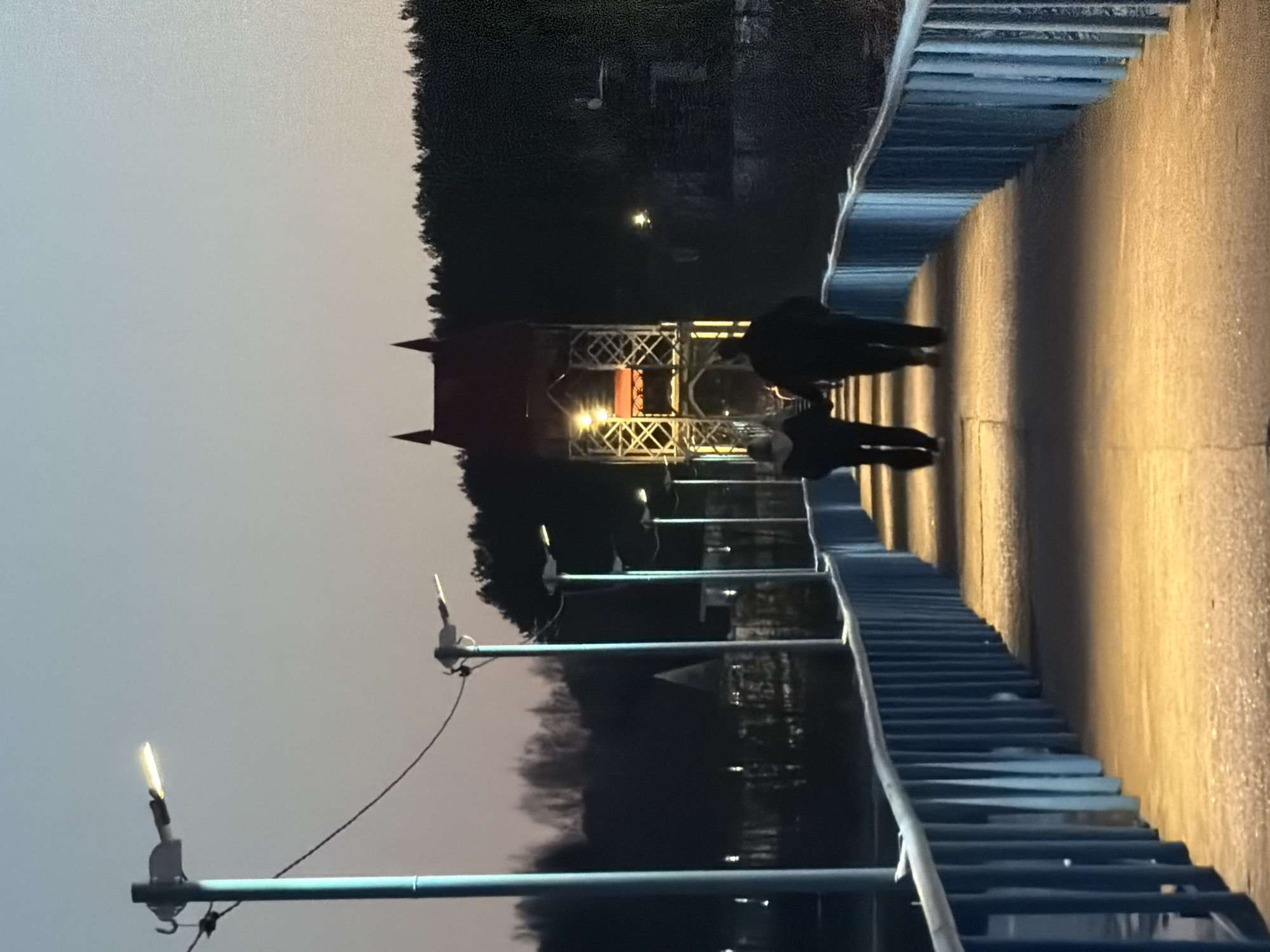 The image captures a scene on a bridge in Izhevsk, Russia, at dusk. Two people are walking on the bridge towards a metal tower illuminated with lights. The bridge has blue railings, and lampposts are spaced along its length. The background features trees and a body of water, likely a river or lake, with some light reflections. The sky is a gradient of light to dark as evening approaches.