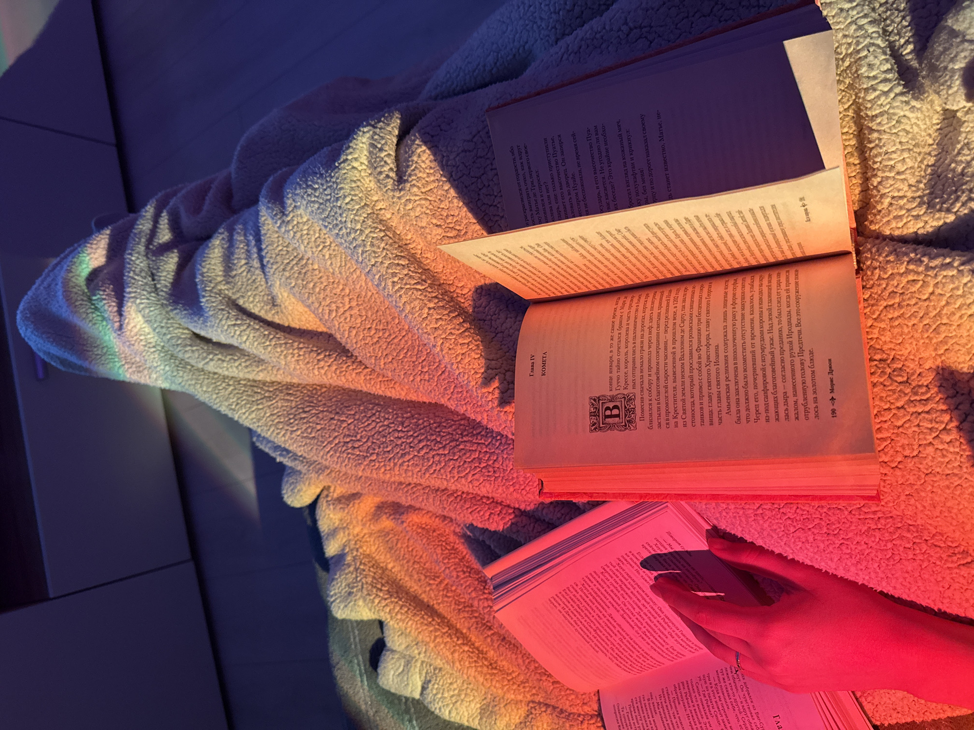 The image captures a cozy indoor scene, likely in the evening or at night, focusing on a reading activity. The primary subjects are a person's hand, several open books, and a light-colored, fluffy blanket or throw.
A hand, illuminated by a reddish light, holds open a book in the lower left portion of the frame. Two other books are prominently open and resting on the blanket in the center and upper right. The blanket itself is draped across a flat, dark surface, possibly a bed or floor, and exhibits folds and a soft texture.
The scene is bathed in vibrant, artificial colored lighting. A rainbow spectrum, with hues of blue, green, and yellow, is visible on the left side of the blanket and the background. A strong red and orange glow illuminates the right side of the blanket, the open books, and the reader's hand, creating a warm and intimate ambiance.
Visible text on the books is in Russian. One book clearly shows "Глава IV КОМЕТА" (Chapter IV COMET) and "190 --- Морис Дрюон" (Maurice Druon), indicating it is a translated work by the French author. Another prominent open book also displays "Глава IV КОМЕТА" and features an ornamental initial "В" on its page. The book held by the hand also contains Russian text and mentions character names such as "Рейчел," "Джек," "Хоуп," and "Синди," along with a place name, "Биг-Сур" (Big Sur), suggesting a novel. The overall impression is one of quiet relaxation and engagement with literature.