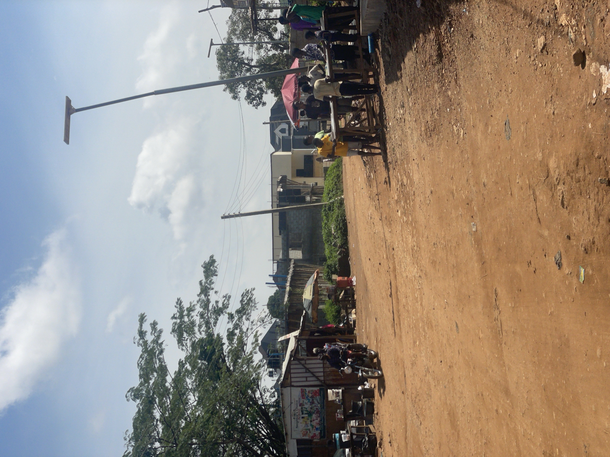 The image depicts a street scene in Kurunduma, Nigeria. A dirt road dominates the foreground, with sparse vegetation and debris scattered across it. On the left side of the road, a small shop with a corrugated metal roof is visible, featuring a sign that reads "Neighbor's Choice." A motorcycle with two people on it is riding past the shop.
Further down the road, a group of young men are gathered around tables, some appearing to be engaged in transactions or conversation. A red umbrella provides shade for some of the individuals. In the background, unfinished buildings and trees are present, along with power lines and streetlights. The sky is partly cloudy, with a bright sun casting shadows. The overall impression is a bustling, informal marketplace or gathering point in a developing urban area.