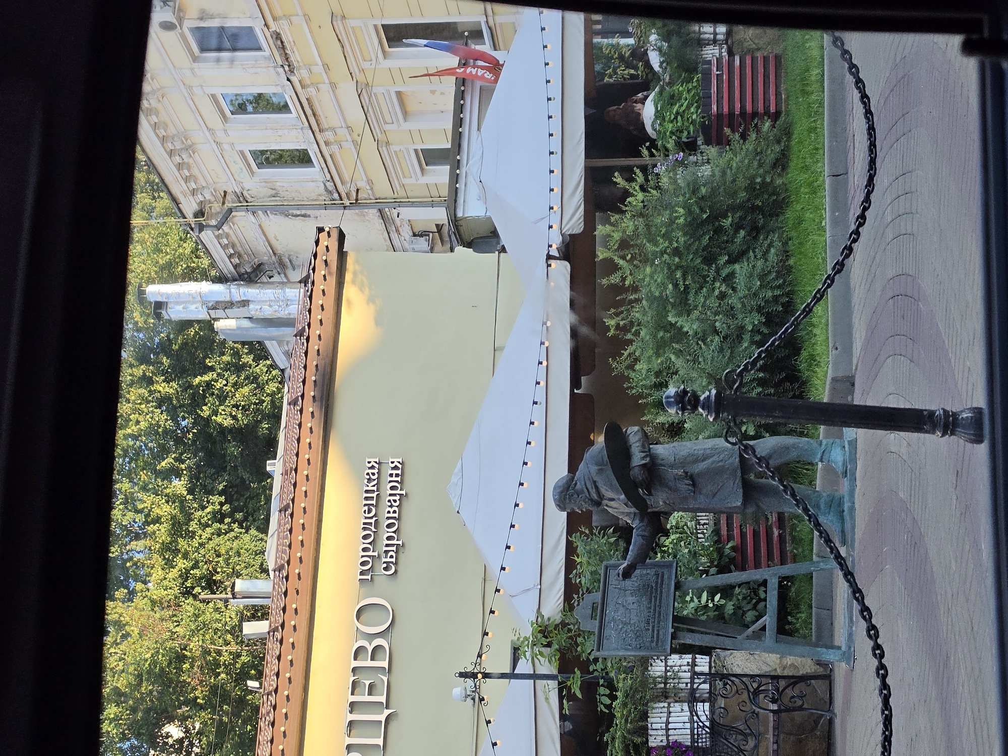 The image depicts a street scene in Nizhny Novgorod, Russia. In the foreground, a bronze statue of an artist stands with a palette in one hand and a brush in the other, facing an easel with a painting. A black chain fence separates the statue area from the sidewalk. Behind the statue, a restaurant with outdoor seating is visible. The restaurant features white umbrellas and a string of lights. The building behind the restaurant is yellow with white trim and several windows. Two Russian flags are hanging from one of the buildings. Trees and greenery are present in the background.