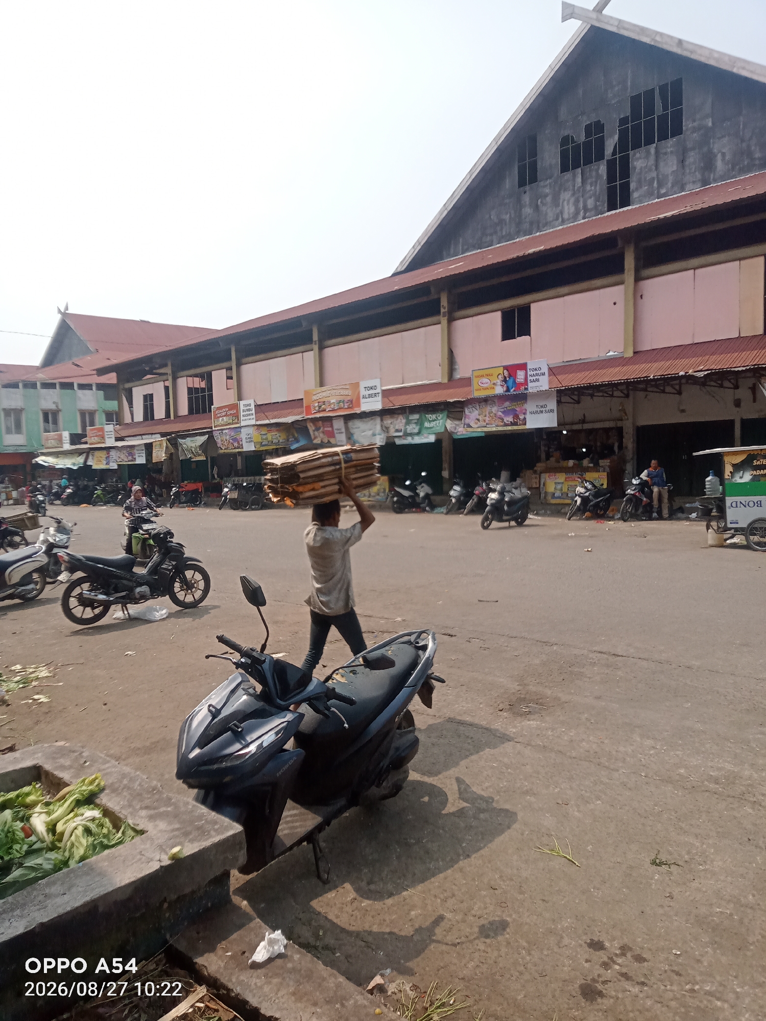 In Jambi City, Indonesia, an outdoor market scene unfolds under a bright sky. A man carries a large bundle of cardboard on his head, walking across a paved area. Several motorcycles are parked around the market, with two in the foreground, one black and one dark gray. The market is lined with stalls and buildings, some of which are decorated with signs in Indonesian. The architecture features red roofs and light-colored walls. In the lower left corner, a concrete structure contains a pile of green leafy vegetables. A water dispenser is visible on a cart near the buildings. The ground is dusty and shows some scattered debris. The image is dated 2026/08/27.