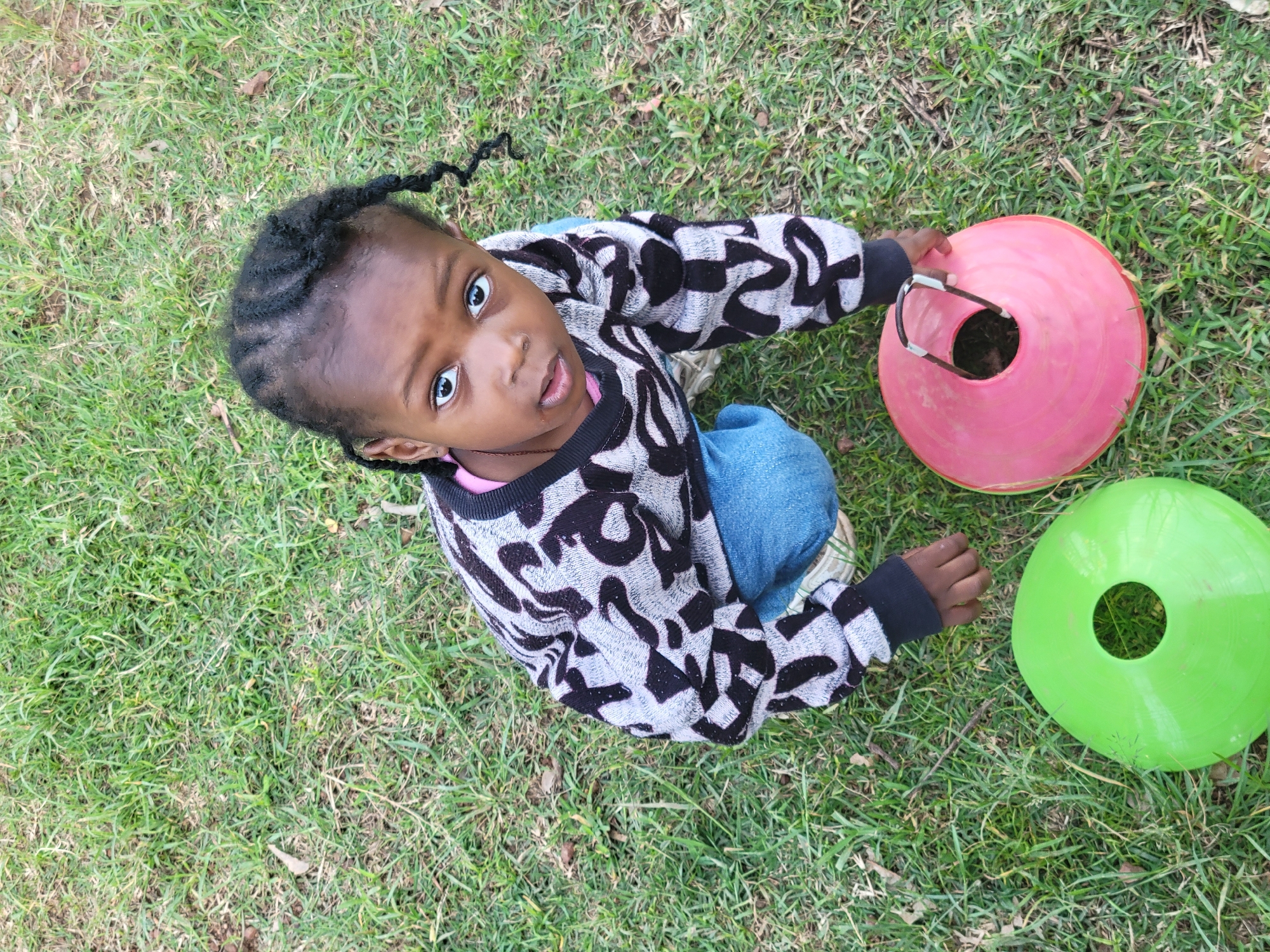 A young Black girl with braided hair kneels on a grassy area, looking up at the camera. She wears a gray sweater with a black and white abstract pattern and blue jeans. In front of her are two traffic cones, one green and one pink. The pink cone has a black handle. The background is a lawn with sparse dry leaves. The overall setting appears to be an outdoor recreational space.