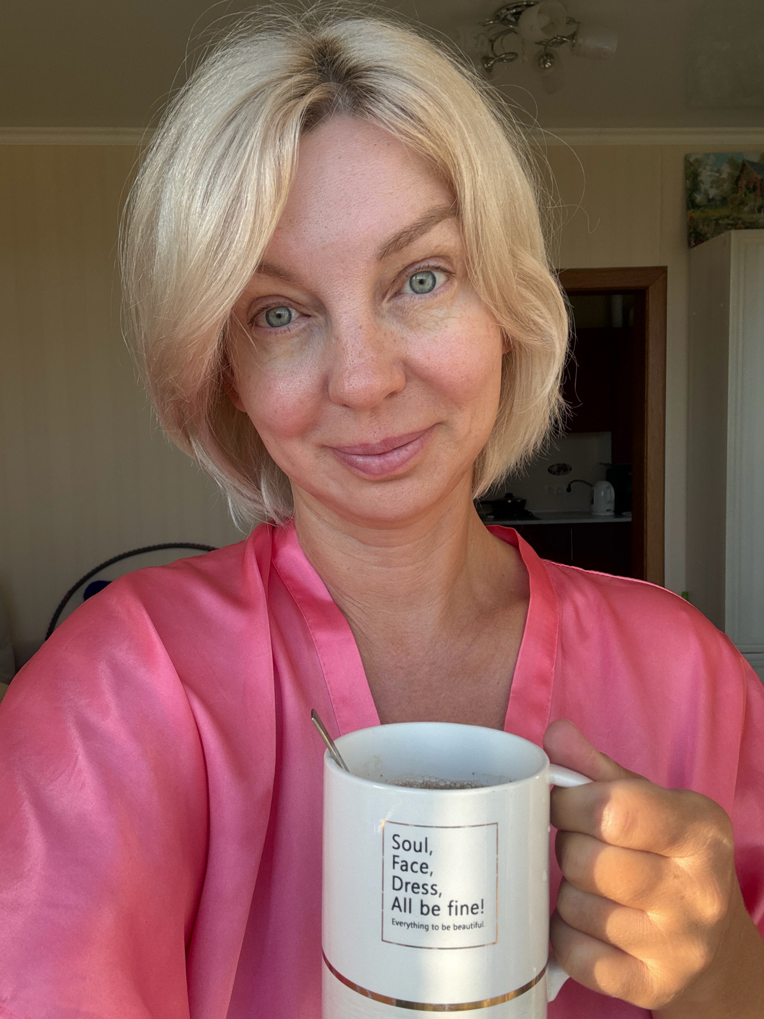 A blonde woman in a pink robe holds a white mug with the text "Soul, Face, Dress, All be fine! Everything to be beautiful." on it. She is indoors, possibly in an apartment, with sunlight streaming in from the right. In the background, there is a doorway leading to another room, a kitchen counter with appliances, and a ceiling light fixture. A painting of a landscape is visible on the wall. The image appears to be a selfie taken in the morning or afternoon. There is no indication of the location being in Sochi, Russia.
