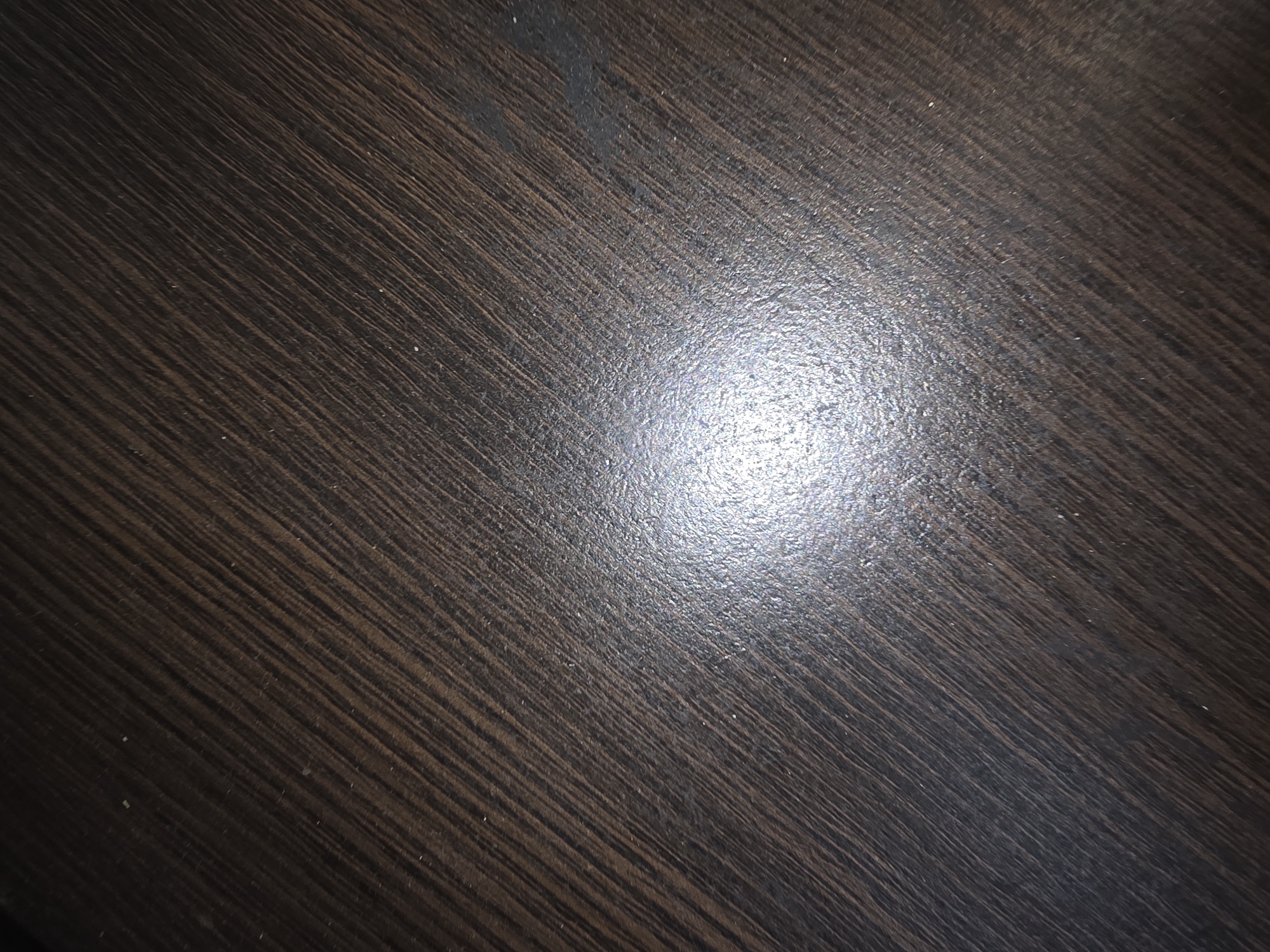 The image shows a close-up, angled view of a dark brown, wood-grained surface, likely a table or countertop. The wood grain runs diagonally from the bottom left to the top right of the frame. A bright, circular reflection of a light source is prominently positioned in the lower-center of the image, indicating a polished or glossy finish. There are a few small, light-colored specks visible on the surface, possibly dust or debris. The overall scene is devoid of any people or specific objects that would indicate a location within Ulan-Ude, Russia, or any other city. The image is purely a textural and lighting study of a common material.