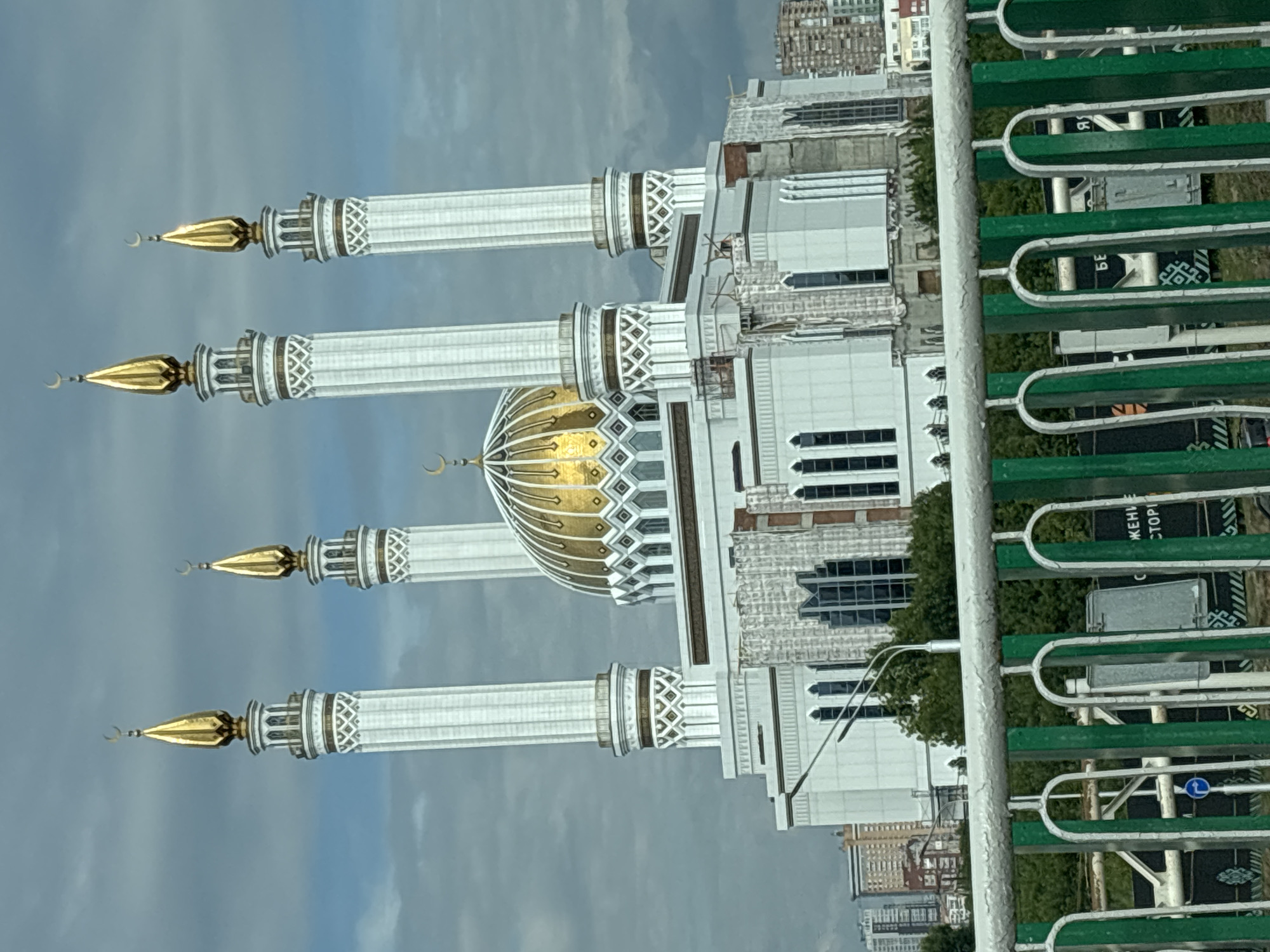 The image shows the Lala Tulpan Mosque in Ufa, Russia. The mosque is a prominent landmark, characterized by its white exterior, golden dome, and four tall minarets topped with golden crescents. The mosque is situated in a city setting, with other buildings and trees visible in the background. A green and white fence is in the foreground, partially obscuring the view of the mosque and the surrounding environment. The sky is overcast with clouds. There are no people visible in the image.