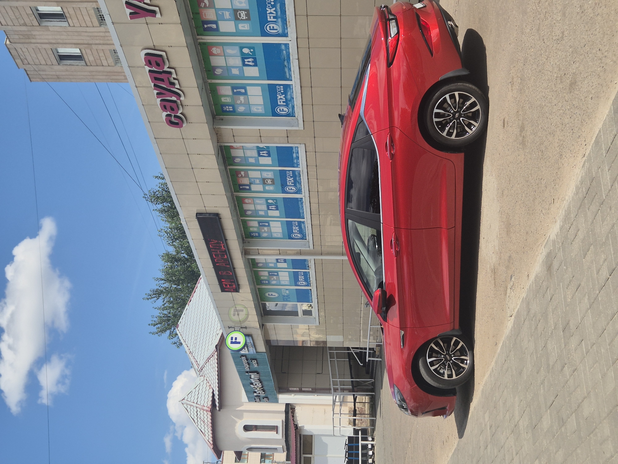 A red sedan is parked on a paved area in front of a commercial building. The building has a light-colored facade with several windows displaying graphics, and prominent signage. One sign reads "сауда" in pink Cyrillic letters. Another section of the building features a green circular logo with the letter "F" in white. A separate structure with a curved, tiled roof is visible to the left of the main building, and a ramp leads to an entrance. Overhead, the sky is blue with scattered white clouds, and power lines are visible. The overall scene suggests an urban commercial area in Kokshetau, Kazakhstan.