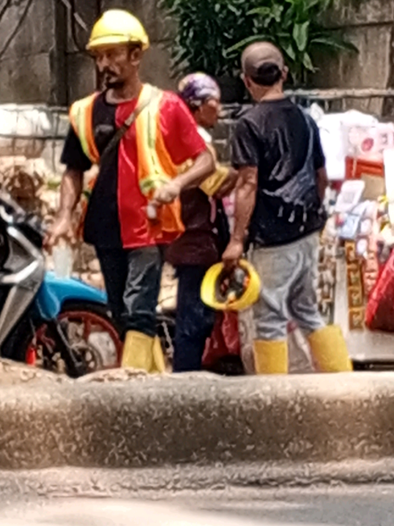 A blurry, medium shot captures three individuals standing on a raised concrete embankment in what appears to be an urban outdoor setting. The person on the left, wearing a yellow hard hat and a red and orange reflective vest over a dark shirt, has a dark mustache and is looking downwards. They are wearing dark pants and bright yellow boots. Beside them, a person in the middle, partially obscured, is wearing a patterned headscarf and a dark top. To the right, another individual, seen from the back, wears a dark t-shirt and light-colored pants, also with bright yellow boots. This person is holding a yellow object, possibly a hard hat or a ring, in their right hand. A motorcycle with a blue body is visible on the left. In the background, there are hints of stalls or vendors with various items, and lush green foliage. The overall impression is a scene of everyday life, possibly a marketplace or street side, in South Tangerang, Indonesia, with construction or labor workers present.