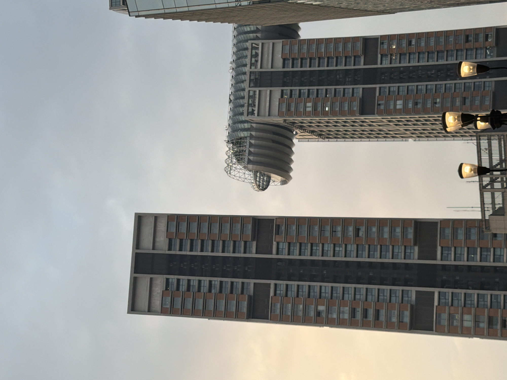 The image displays a street-level perspective of three modern skyscrapers in the Yuzhong District, Chongqing, China, under an overcast sky showing a warm, yellowish light on the lower left, indicating either dawn or dusk. On the left, a tall, rectangular residential or office building features a facade with numerous repeating rectangular windows set into brown-orange panels, interspersed with darker grey vertical and horizontal structural elements. To its right, another skyscraper, part of the Raffles City Chongqing complex, is topped by a prominent, elongated sky bridge that connects to an adjacent, unseen structure. This sky bridge exhibits a complex, curvilinear glass-domed roof and a ribbed, solid base. A third, darker, glass-clad building is partially visible on the far right, bordering the skyscraper with the sky bridge. In the lower foreground, several streetlights with illuminated, amber-colored bulbs are visible, along with dark metallic structural elements. No individuals or specific actions are depicted in the scene.