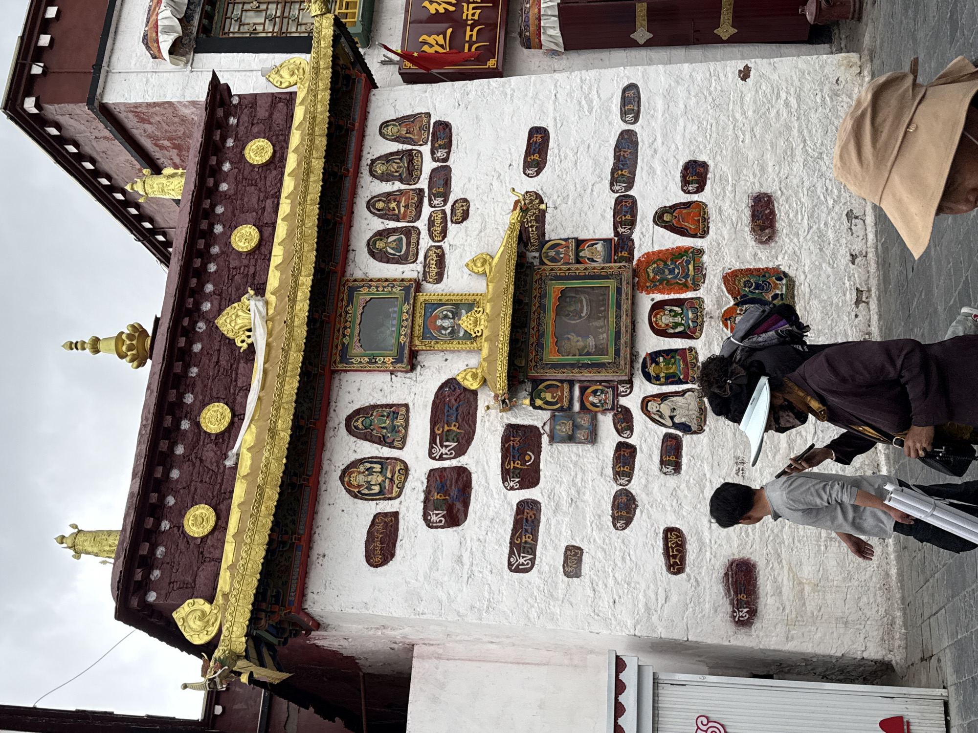 The image captures a scene in Chengguan District, Lhasa, Tibet Autonomous Region, China. The subject is an exterior wall of a religious building adorned with a combination of framed artwork and painted Tibetan scripts or religious images.
The building features a decorative, multi-tiered roof structure with a burgundy color and golden accents. The wall is painted white and displays various framed pictures, as well as painted artworks.
Three people are visible near the wall. One person is wearing a tan hat. Another person appears to be holding a pen or pointing at the wall. A third person is holding rolled-up papers. The interactions between them seem to indicate they are either studying, preserving, or documenting the artwork on the wall.
The environment suggests an urban setting within a historically or culturally significant area, due to the style of the architecture. The sky is overcast, indicating possibly an outdoor setting.