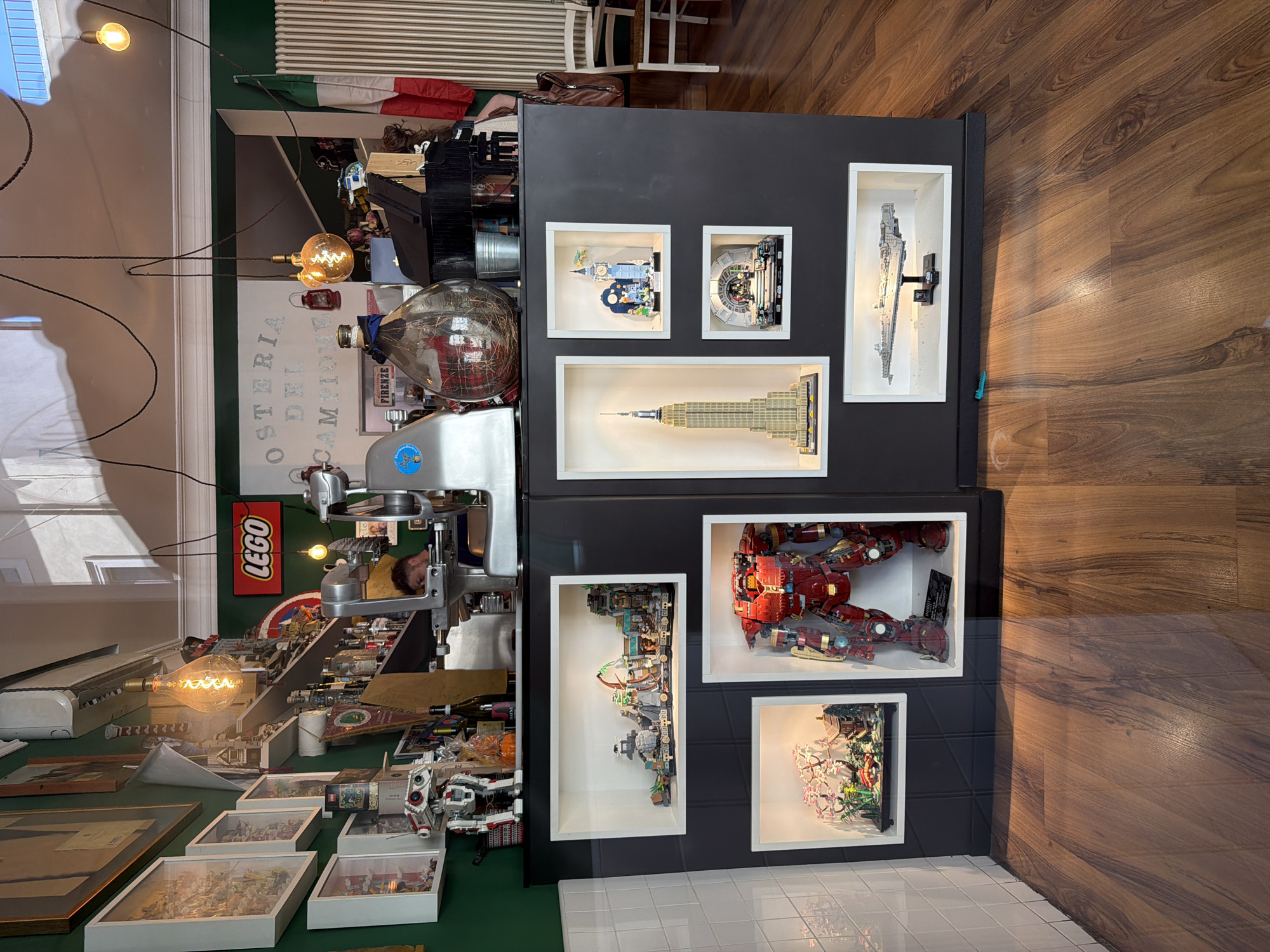 The image shows a complex and cluttered interior scene, likely a shop or restaurant, in Mantua, Italy. The foreground features a wooden floor and a white tiled wall. A dark shelving unit displays Lego models in white frames. Above the shelving unit is a chaotic space containing a meat slicer, various objects, and a Lego sign. Lightbulbs hang from above, casting a warm glow. A large window offers a partial view of the interior. The name "OSTERIA DEL CAMPIONE" is visible on the wall, confirming the location in Mantua. The scene appears to be a unique blend of culinary preparation and Lego collection display.