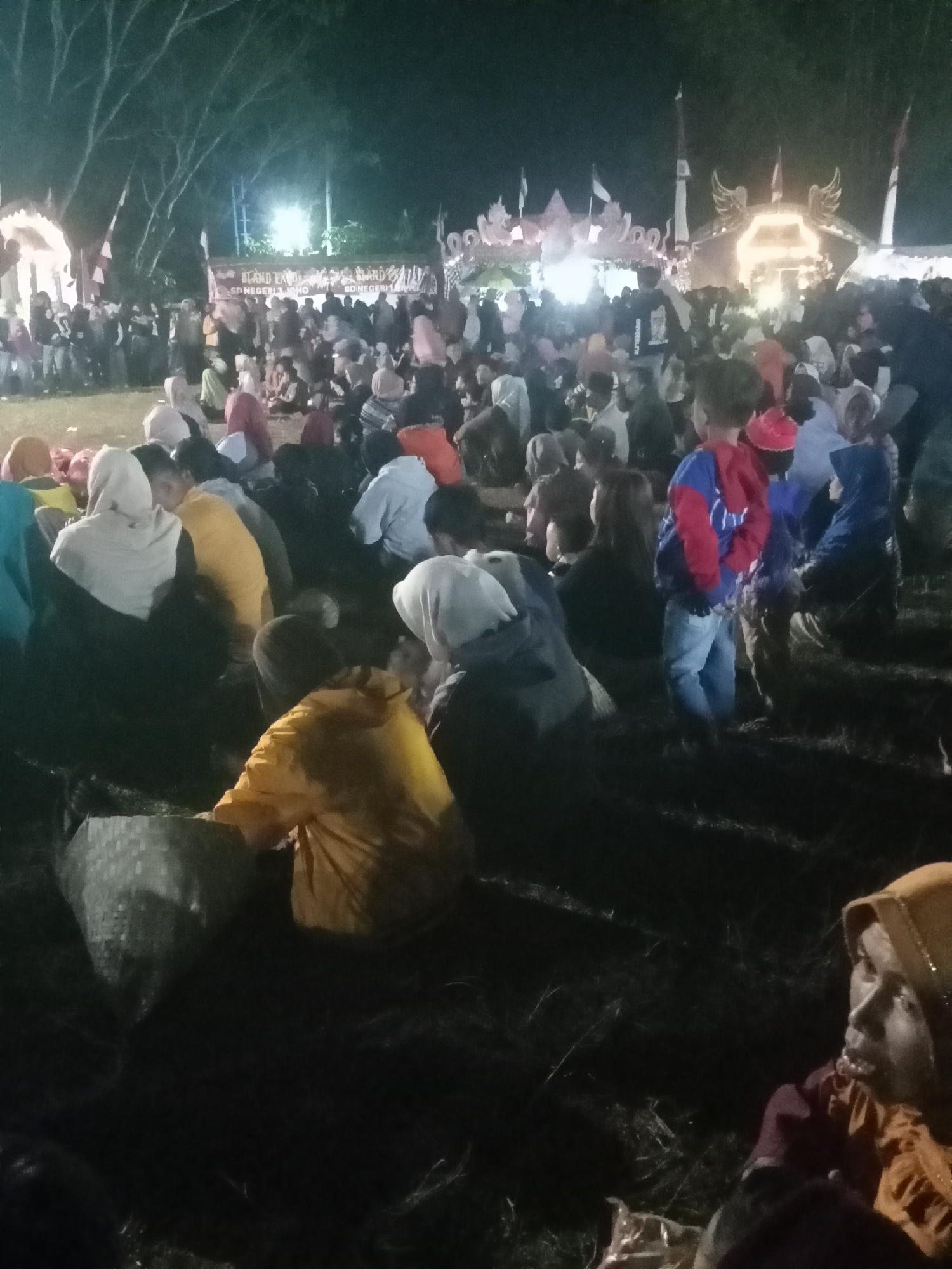 The image depicts a nighttime outdoor gathering in Pule, Indonesia, possibly a festival or event. A large crowd of people, many wearing headscarves, are seated on the ground. In the background, there are illuminated structures and banners, suggesting a decorated venue. Several children are visible in the foreground, some standing and others sitting. The ground appears to be covered in dry grass or hay. The lighting is dim, with bright lights originating from the structures in the background.