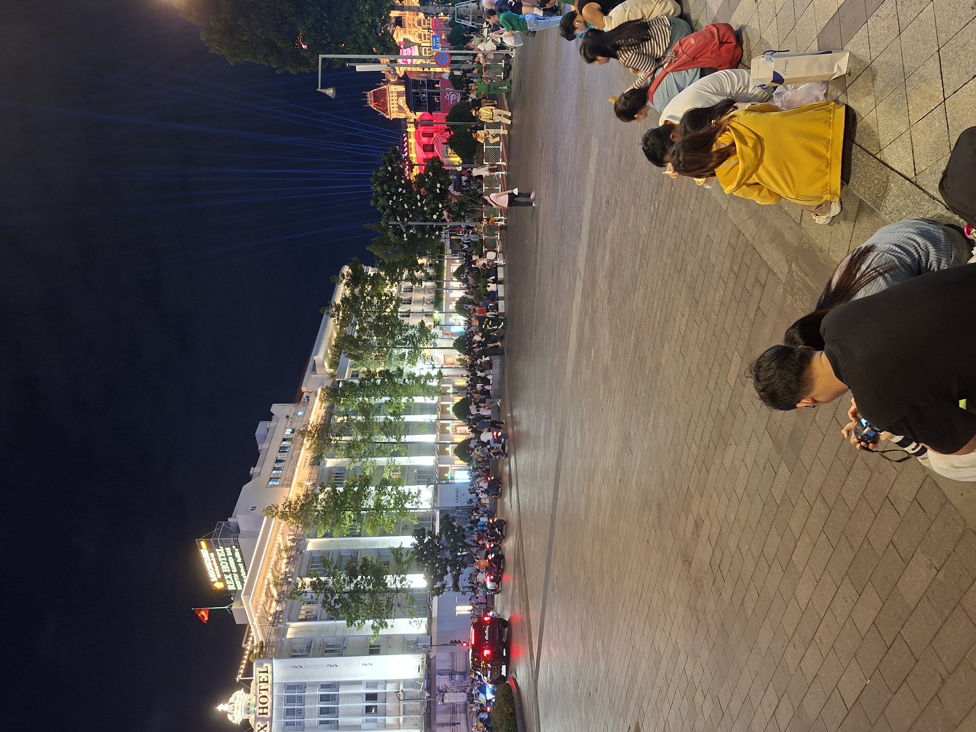 The image captures a nighttime scene in Thủ Đức, Vietnam, likely a public square or pedestrian area in front of the Rex Hotel. The Rex Hotel, a prominent white building with illuminated signage and decorative lights, dominates the background. A flagpole with a Vietnamese flag is visible on its rooftop. In the foreground, a group of people are seated on the ground, facing away from the camera, some appearing to be using phones or cameras. Another person in the foreground is holding a camera and looking at it. A paved walkway with a brick pattern occupies the central area. Beyond the seated individuals, a street scene unfolds with traffic, including cars and motorcycles, and a crowd of people gathered. Illuminated buildings and a display of bright blue light beams emanating from the sky are visible in the distance. The overall atmosphere is lively and suggests a public event or gathering.