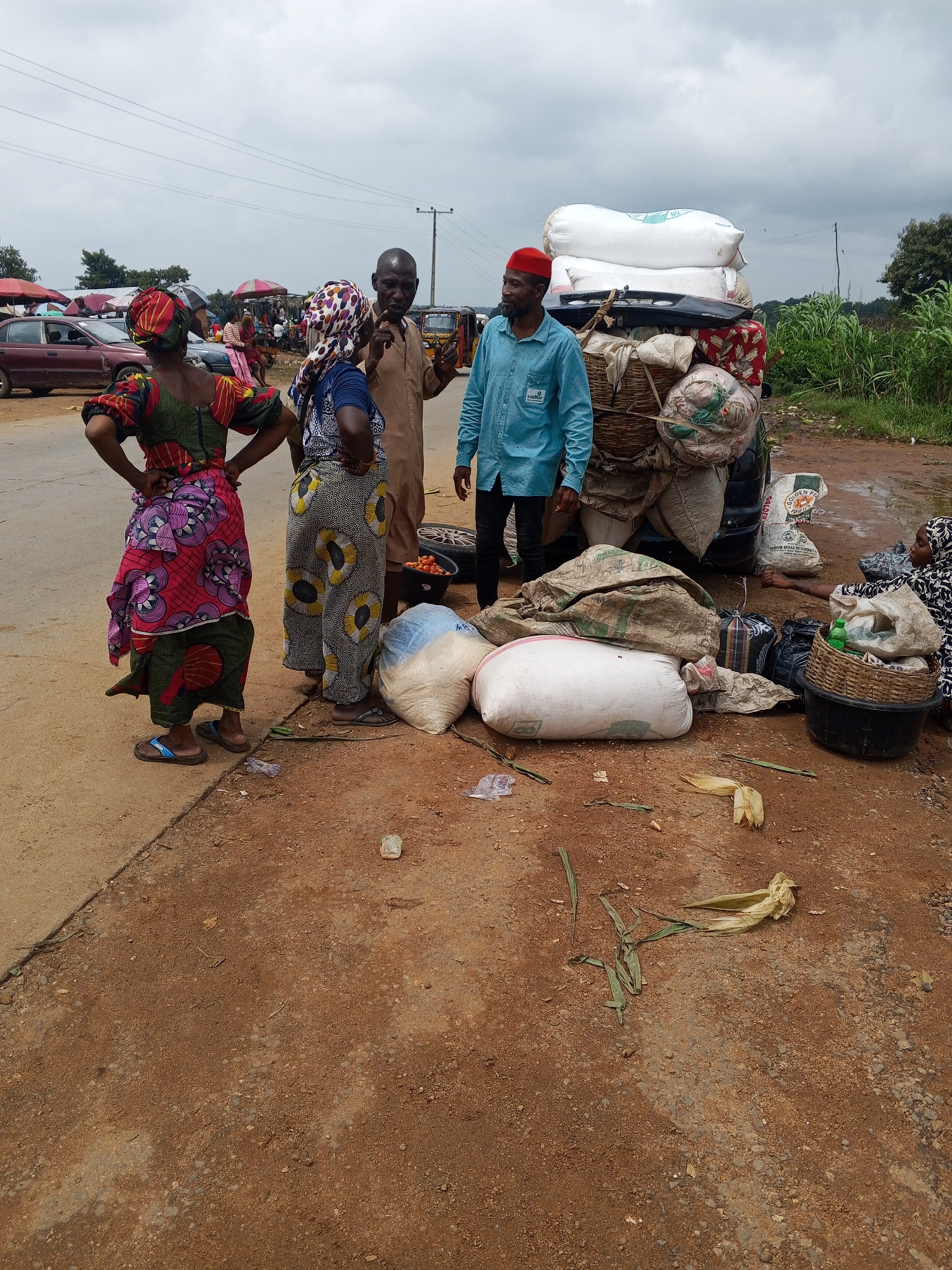 In Oduape, Nigeria, a group of people are gathered by a roadside market. Several individuals are standing near a vehicle overloaded with various goods, including large sacks of produce and what appear to be personal belongings. One woman in a colorful patterned dress stands with her hands on her hips, facing away from the camera. Another woman in a headscarf and patterned skirt is talking to a man in a brown tunic. A man wearing a blue shirt and a red cap is standing next to the loaded vehicle, looking to the left. In the foreground, there are more large sacks, some appearing to be filled with grains or flour, and a woven basket. The ground is a mix of dirt and a paved surface, with some debris scattered around. In the background, more market stalls with umbrellas are visible, along with parked cars and lush green vegetation. The sky is overcast.