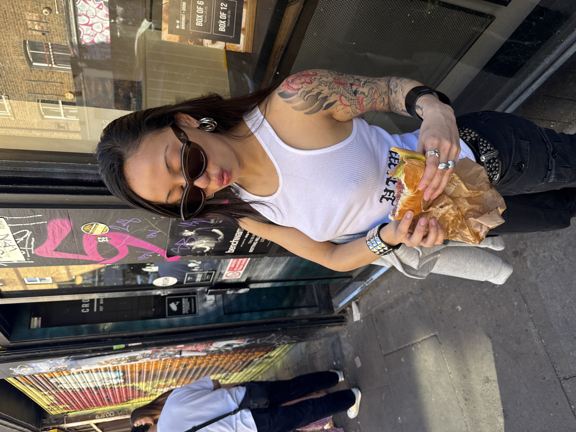 A street scene features a woman with a tattoo on her arm holding a sandwich in a brown paper bag. She has dark hair, wears sunglasses, a white ribbed tank top, and black pants with a studded belt. She also sports multiple rings and ear piercings. The environment includes a shop window with signage mentioning "DOUGHNUT + DRINK" and "BOX OF 6" / "BOX OF 12" along with prices. On the storefront next to the window, there is graffiti and stickers. A sign also says "CRO 157 BR". In the foreground, a partial view of another individual wearing a white top and black pants can be seen. The setting is a street, identifiable by the pavement underfoot. According to the signage, the location is likely in Notting Hill, London.
