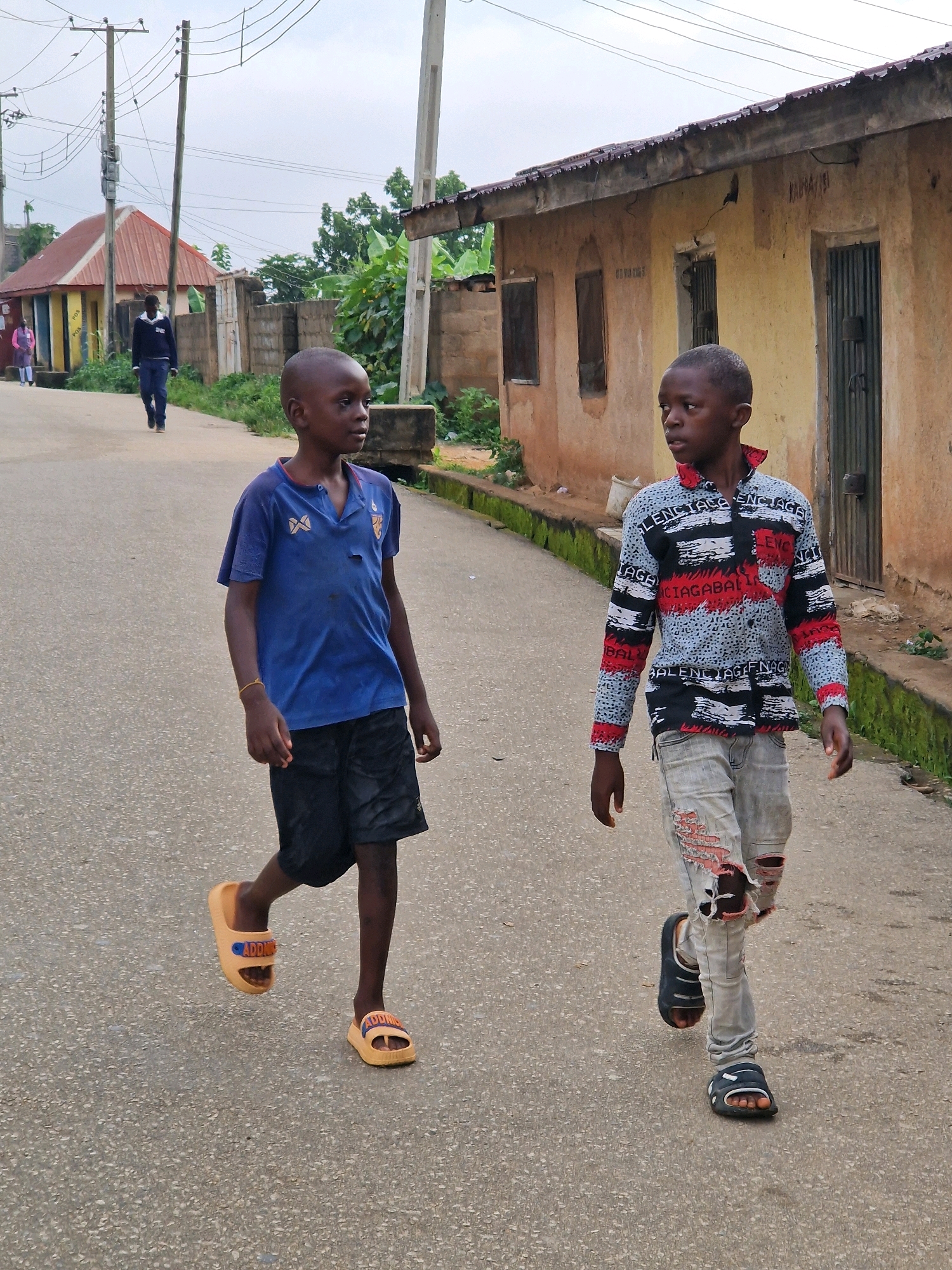 Two young boys walk on a paved road in front of buildings in Jos, Nigeria. The boy on the left wears a blue polo shirt and black shorts. He has an orange sandal on his left foot and an orange sandal on his right foot. The boy on the right wears a patterned long-sleeved shirt and ripped light-colored jeans. He has a dark sandal on his left foot and a dark sandal on his right foot. The buildings are made of earth-toned material with corrugated metal roofs. Electrical poles and wires are visible in the background. The sky is overcast.