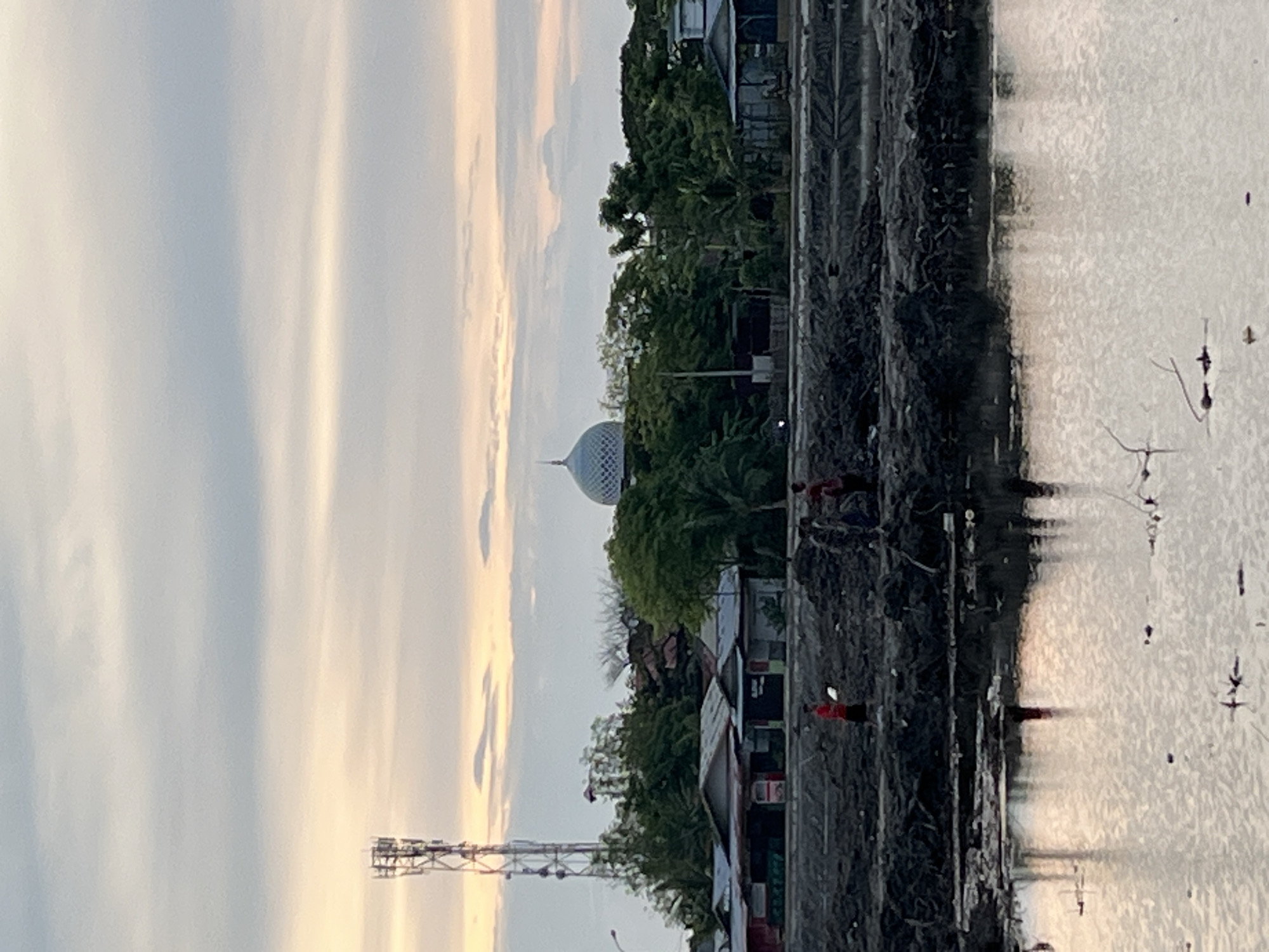 The image depicts a scene in Lhokseumawe, Indonesia, featuring a waterfront area with muddy banks. In the foreground, a body of water reflects the sky and surrounding structures. The middle ground shows a raised, dark, muddy embankment where several individuals are present. One person, wearing a red shirt, stands alone, holding a white object. Further to the right, a group of people, some appearing to be children, are gathered. Behind them, a row of modest buildings with corrugated metal roofs stretches across the landscape. Dominating the background are lush green trees and a prominent mosque with a silver dome. To the left of the mosque, a tall telecommunications tower stands against a pale, overcast sky with hints of sunset colors. The overall impression is of a functional, possibly developing, urban environment near the water.