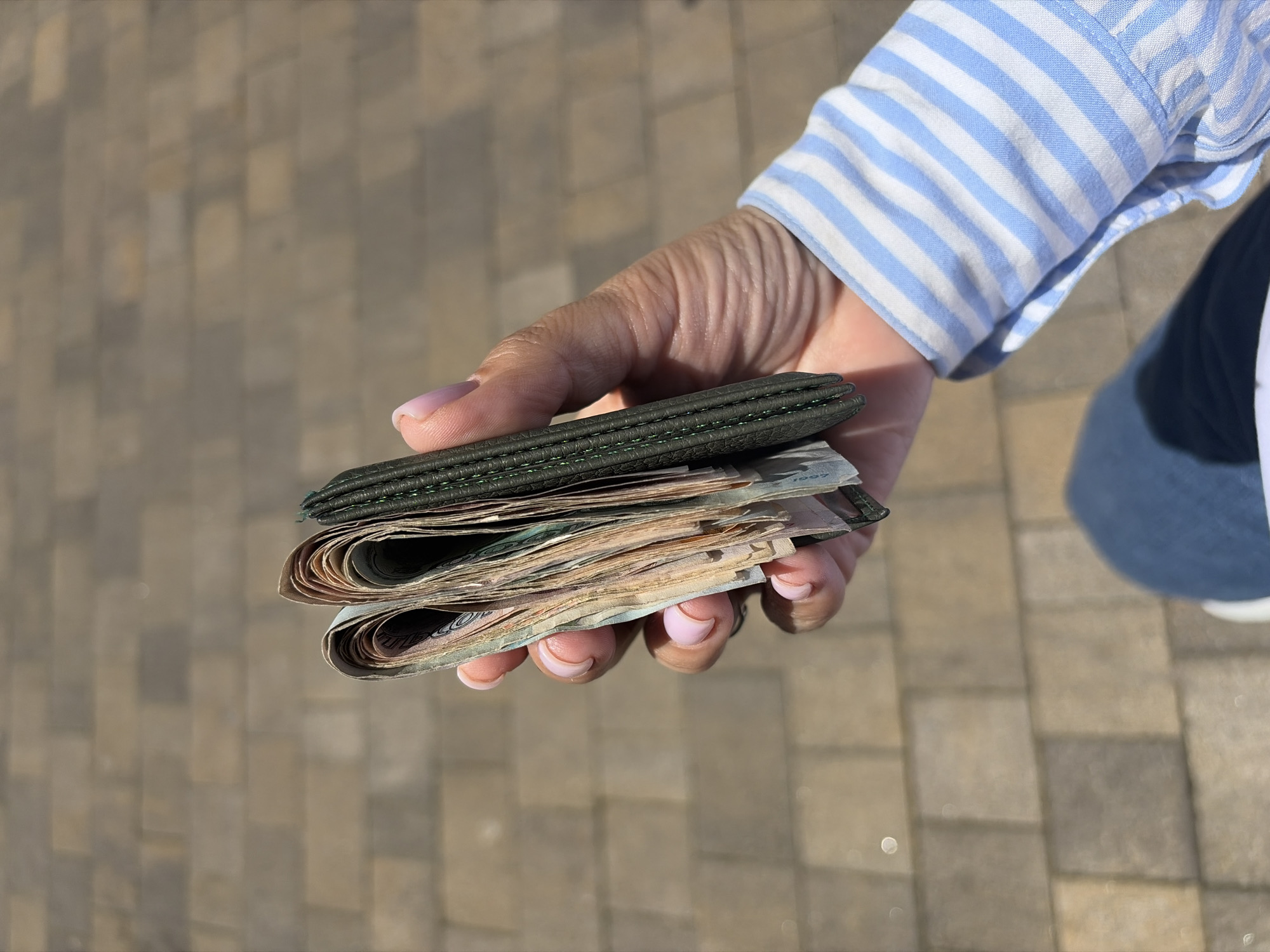 A close-up shot captures a person's hand holding a thick stack of banknotes and a dark green, textured wallet or money clip. The hand, with light pink French-manicured fingernails, is positioned over a light-colored paved ground, likely cobblestones or patterned tiles, indicating an outdoor setting in bright daylight. The person is wearing a blue and white striped long-sleeved shirt, visible around the wrist, and a partial view of blue jeans or pants is seen in the lower right background. The banknotes appear to be Russian Rubles, consistent with the Sochi, Russia location context, and are slightly curled, showing signs of use. The year "1997" is faintly visible on one of the banknotes. The scene suggests a person handling cash outdoors on a sunny day.