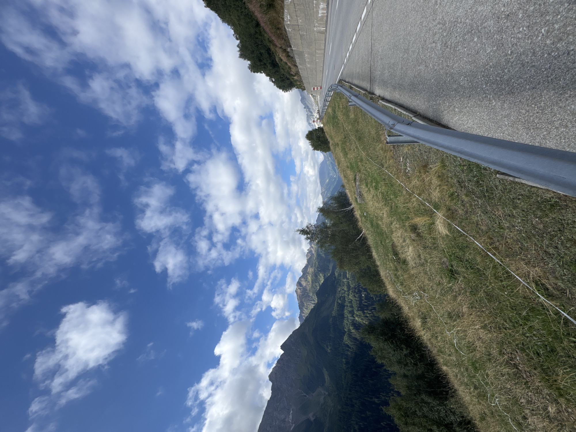 The image depicts a scenic mountain road in Airolo, Switzerland, under a bright blue sky with scattered white clouds. The road curves to the right and ascends along a steep, grassy mountainside. A grey metal guardrail runs along the edge of the road, separating it from the slope. The mountains in the distance are covered in dense green forest, with jagged peaks rising towards the sky. The foreground shows dry, golden grass and a thin, white wire fence. The overall impression is one of a beautiful, remote, and potentially challenging mountain pass.