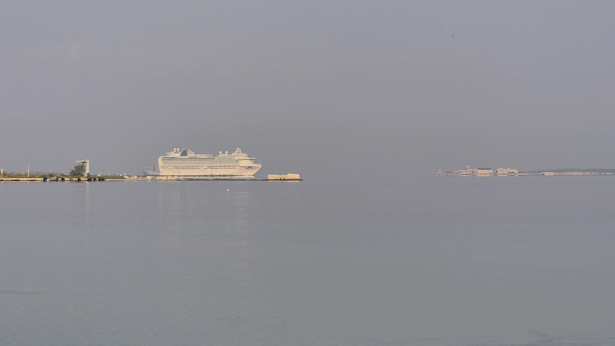A wide-angle view captures the calm, gray waters of the Mar Piccolo bay in Taranto, Italy. A large, white cruise ship with red and blue accents is docked on the left side of the frame. A dock with a crane and some smaller boats is also visible near the cruise ship. In the distance, on the right side, a cluster of buildings sits on a narrow strip of land, with a red lighthouse in the foreground. The sky is a hazy, light gray, suggesting a slightly overcast or foggy day. The water is flat and reflective, mirroring the muted colors of the sky and the distant structures.