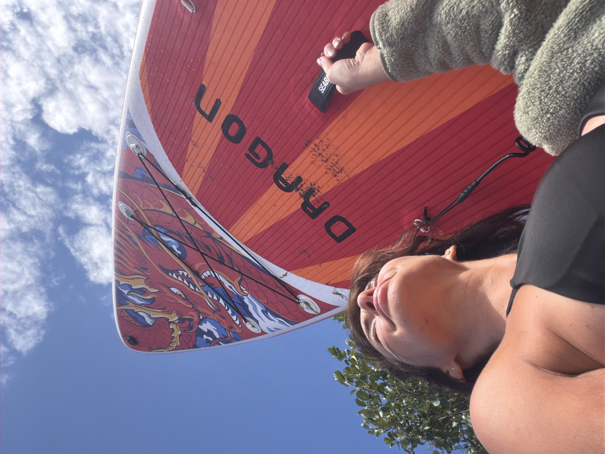 A low-angle shot captures a woman in a black bikini top, with her face tilted upwards and eyes closed, seemingly enjoying the sun. She is partially obscured by a red and orange paddleboard adorned with a blue and red dragon design and the word "DRAGON" in black lettering. Her right hand, wearing a fuzzy green sleeve, is holding onto a black handle attached to the paddleboard with the word "SEAB" visible. To the left, a portion of a green leafy tree is visible. The background is a clear blue sky with scattered white clouds. The scene appears to be outdoors, likely near water, given the presence of the paddleboard. The location is not specified as being in Vyazy, Russia.