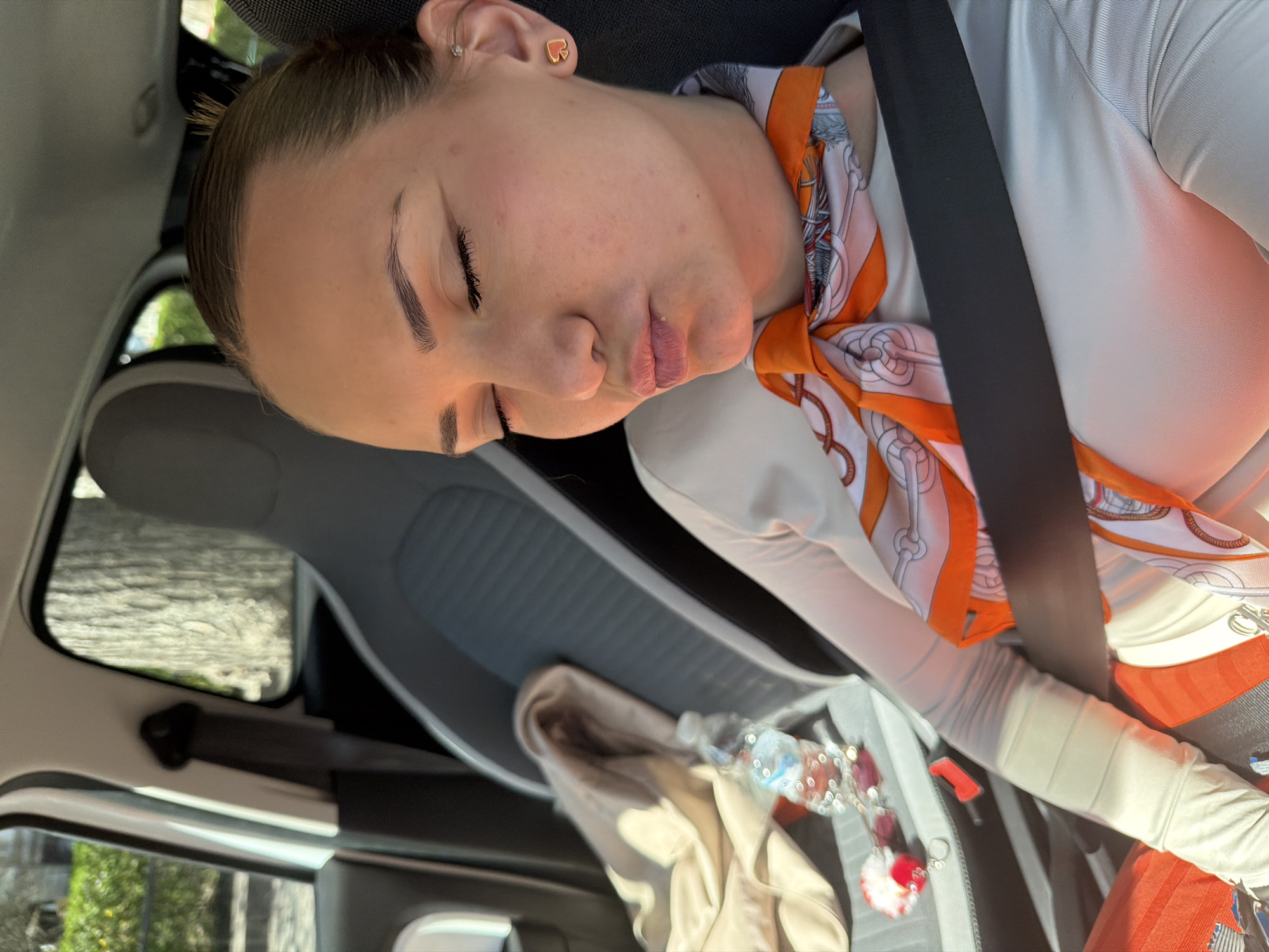 A medium shot depicts a young woman sleeping in the passenger seat of a car. She is wearing a white long-sleeved shirt and an orange and white patterned scarf around her neck, secured by a black seatbelt. Her head is tilted to the side. In the background, the car's interior is visible, including the passenger seat and items like a beige jacket, a water bottle, and a decorative trinket hanging from the rearview mirror. The car's window shows an out-of-focus scene of greenery, suggesting an outdoor setting, consistent with a city like Sofia, Bulgaria.