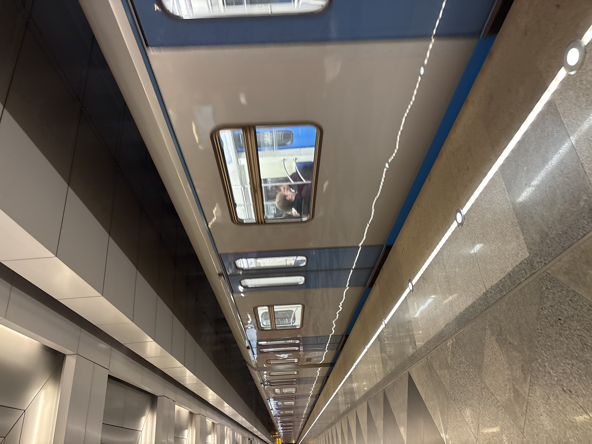 A low-angle shot depicts the interior of the Vnukovo Metro station in Moscow, Russia, with a focus on the train's ceiling and windows. The train's ceiling is white with blue trim, and a series of windows reflect the interior of another train car, revealing passengers inside. The station's architecture is visible on either side of the train, featuring modern, geometric wall panels. Recessed lighting fixtures run parallel to the train, illuminating the station. The image suggests a bustling urban transportation environment.