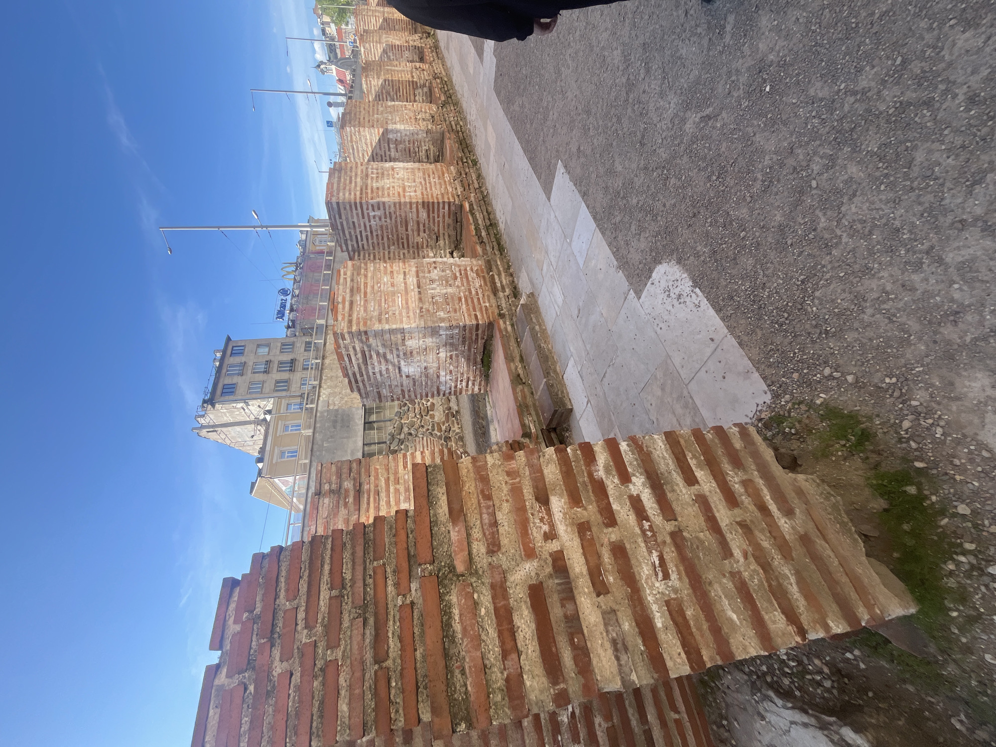 The image, taken outdoors on a clear day, depicts ancient brick structures juxtaposed with a modern urban setting. A series of reddish-brown brick ruins, resembling rectangular foundations or pillars, line the left side of the frame. These structures are made of elongated bricks with visible mortar, showing signs of age and historical significance.
Adjacent to these ruins, a paved path with light-colored tiles transitions into a gravelly, dirt-covered area. A person's lower torso and legs, dressed in dark attire, are partially visible in the upper right portion of the frame, suggesting their presence near the walkway.
In the background, under a bright, clear blue sky with minimal clouds, modern city buildings rise. These multi-story structures feature numerous windows and contemporary architecture. Tall, slender streetlights are interspersed throughout the scene.
Visible text and logos on the buildings in the background include the "Allianz" name and emblem, and the distinctive golden arches logo of "McDonald's." The scene is brightly lit by natural sunlight, indicating daytime. The combination of ancient ruins and modern commercial buildings points to an archaeological site integrated into a bustling city environment, consistent with locations like Sofia, Bulgaria.