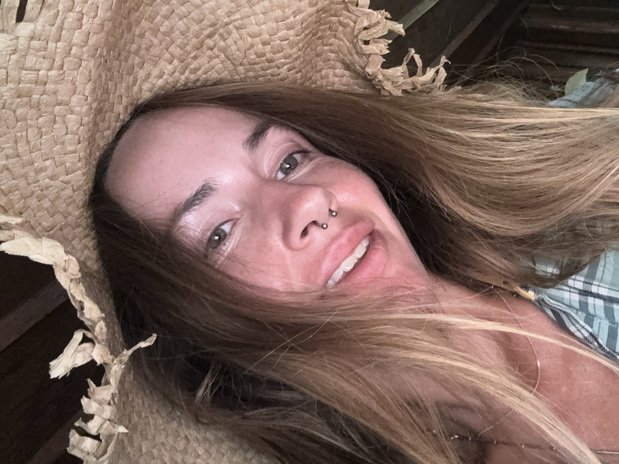 A close-up selfie shows a woman wearing a wide-brimmed straw hat with frayed edges. Her long, light brown hair is visible around her face. She has green eyes, a septum piercing, and is smiling with her teeth showing. She is wearing a plaid shirt and a delicate necklace. The background is dark and appears to be an indoor space with wooden elements. The lighting suggests the photo was taken in natural light.