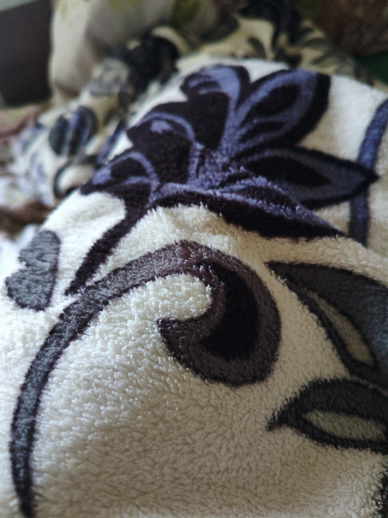 The image shows a close-up, slightly blurred view of a fuzzy, white blanket with a dark blue floral pattern. The blanket is folded and draped, creating soft curves and shadows. The floral design consists of stylized leaves and petal-like shapes. The lighting is diffused, highlighting the texture of the fabric. The location in Rîșcani, Moldova, is not visually represented in the image itself.