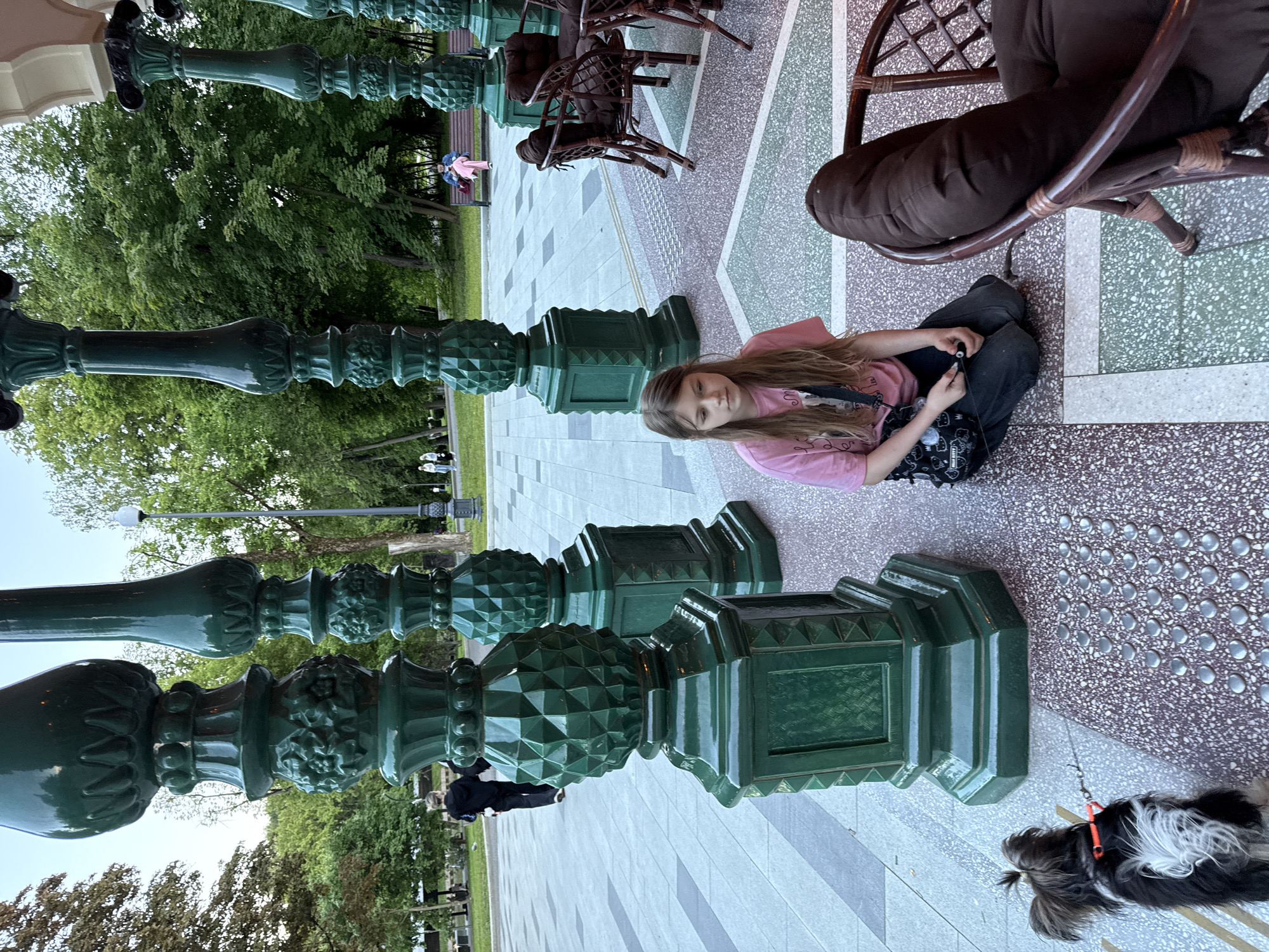 This image, when viewed upright, captures an outdoor scene in a public park or square in Moscow, Russia.
**Subjects:**
In the foreground, a young girl with long, light brown hair is seated on the patterned pavement. She wears a pink long-sleeved shirt and dark pants, looking calmly towards the camera while holding a small black object, likely a phone. To her right, an adult is partially visible, seated at a table or chair, wearing dark clothing. A small, shaggy black and white dog is lying on the pavement near the girl, attached to a red leash. In the background, several other people can be seen walking or sitting amidst the greenery.
**Objects:**
Prominently featured throughout the scene are large, decorative green posts. These ornate structures, with intricate carvings and faceted designs, resemble oversized chess pieces or elaborate bollards, lining the paved area. A dark wicker chair and part of a table are visible on the right side, occupied by the adult. The girl has a dark bag or purse with white patterns beside her. A lamppost is discernible in the far background.
**Scene and Setting:**
The setting is a well-maintained outdoor pedestrian area, characterized by a diverse patterned pavement featuring sections of grey, white, and speckled dark red/brown, interspersed with green tiled areas. A tactile paving strip with circular bumps is also present. Beyond the paved zone lies a lush green park with numerous mature trees, indicating a verdant urban space. The overall ambiance is that of a relaxed public gathering spot.
**Activity:**
The young girl appears to be resting or engrossed with her device, while the adult companion is seated nearby. The dog is also at rest. People in the background are casually enjoying the park environment.
**Notable Details:**
The time of day appears to be daytime, possibly late afternoon or early evening, with soft, clear light. The weather is fair. The distinctive green decorative posts are a key visual element of this Moscow location. No visible text is legible in the image.