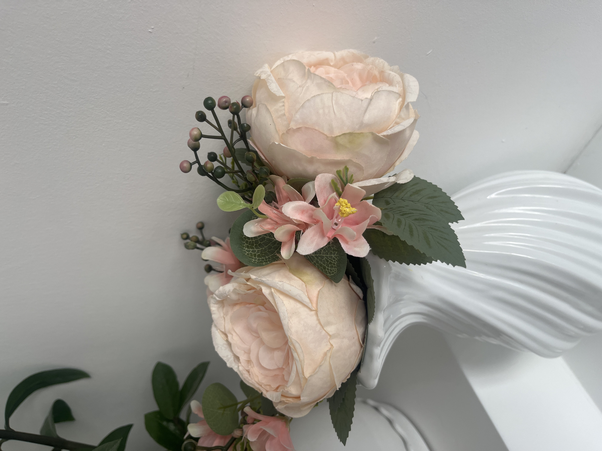 The image shows two peach-colored roses with green leaves and small green berries, arranged against a white background. The roses appear to be artificial. A white, sculpted object is partially visible on the right side of the image. Part of another arrangement of greenery is seen at the bottom left. No people or actions are visible. The location in Bac Nha Trang Ward, Vietnam is not evident from the visual content of the image.