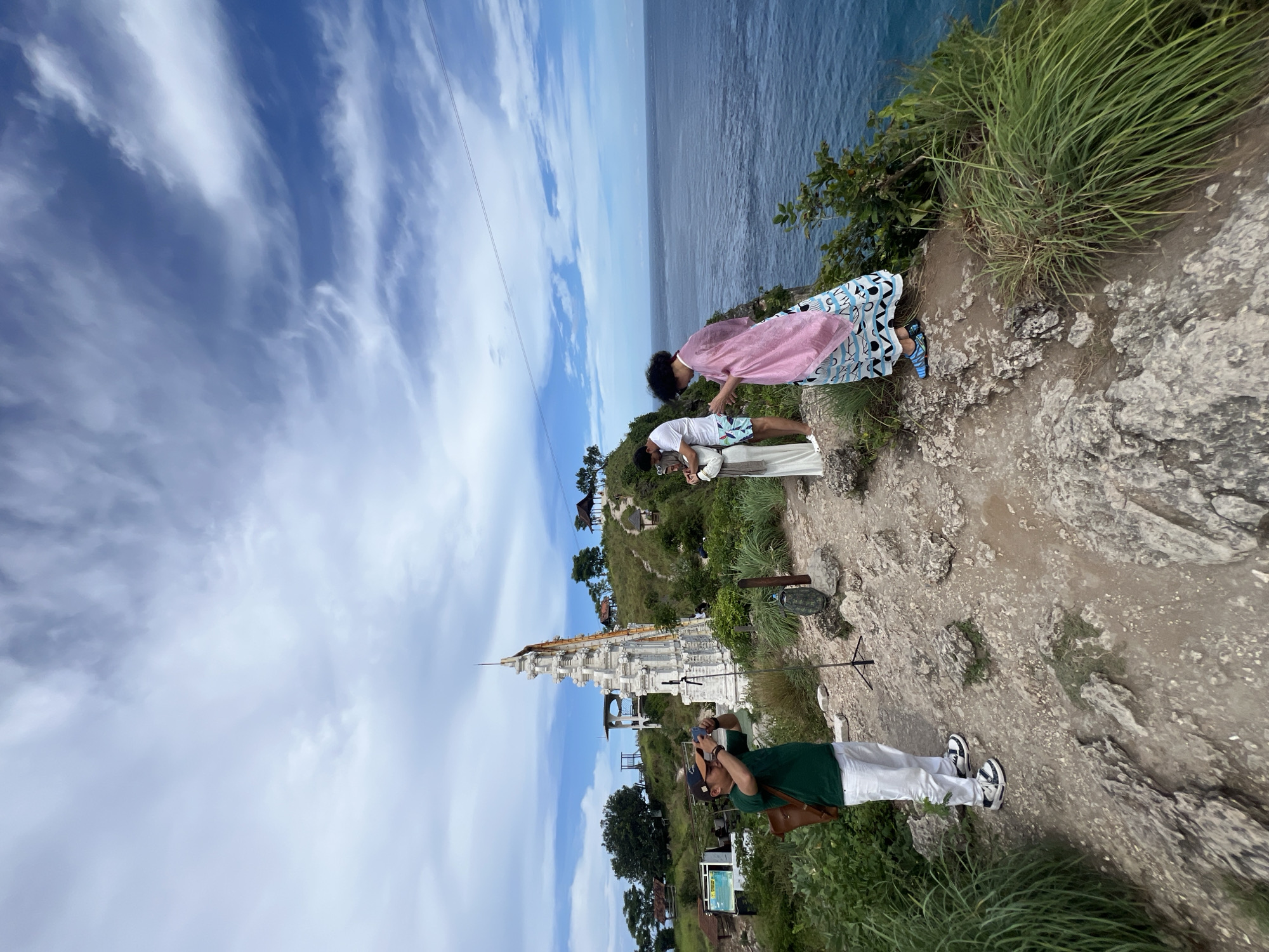 This image depicts a scenic coastal view from a cliffside in Nusa Penida, Indonesia, under a bright, sunny sky with scattered wispy clouds. The deep blue ocean stretches to the horizon, meeting the sky.
Three individuals are visible in the scene. In the foreground, near the cliff's edge, one person wears a pink sheer top over a patterned blue and white skirt or sarong and blue sandals, looking out towards the sea. Adjacent to them, a second person in a white t-shirt and patterned shorts leans over a low stone barrier, also observing the ocean. The cliff features rugged terrain with patches of lush green grass and bushes.
Further back and to the left, a man in a green polo shirt, white trousers, and a blue cap holds a smartphone up, appearing to take a photograph. He wears black and white sneakers and carries a brown shoulder bag. Behind him stands a distinctive white temple or multi-tiered structure with spires, indicative of local architecture, situated on a verdant hillside.
The weather appears clear and pleasant. Utility lines are faintly visible against the sky. A small sign with unreadable text is present near the base of the temple structure. The overall impression is one of enjoying a picturesque natural landscape and cultural landmark.