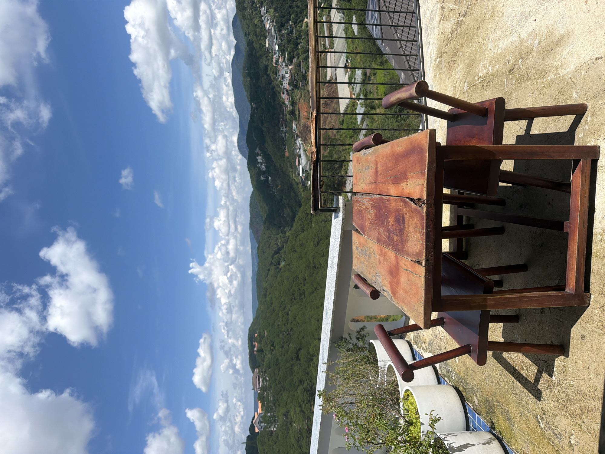 The image depicts an outdoor dining set with a rustic wooden table and four chairs positioned on a concrete patio. The table is rectangular with a weathered, distressed wood finish. Two chairs are visible on the left side of the table and two on the right. The chairs are made of dark wood and have a simple, backless design with curved armrests.
In the background, a lush green valley stretches out, dotted with trees and houses. The landscape rises into rolling hills and mountains under a bright blue sky filled with fluffy white clouds. A white railing runs along the edge of the patio, separating it from the valley below. Several white planters with greenery are also visible on the left side of the patio. A black metal fence runs parallel to the railing, further enclosing the patio area. The scene suggests a viewpoint overlooking a residential area nestled within the natural landscape of Da Lat, Vietnam.