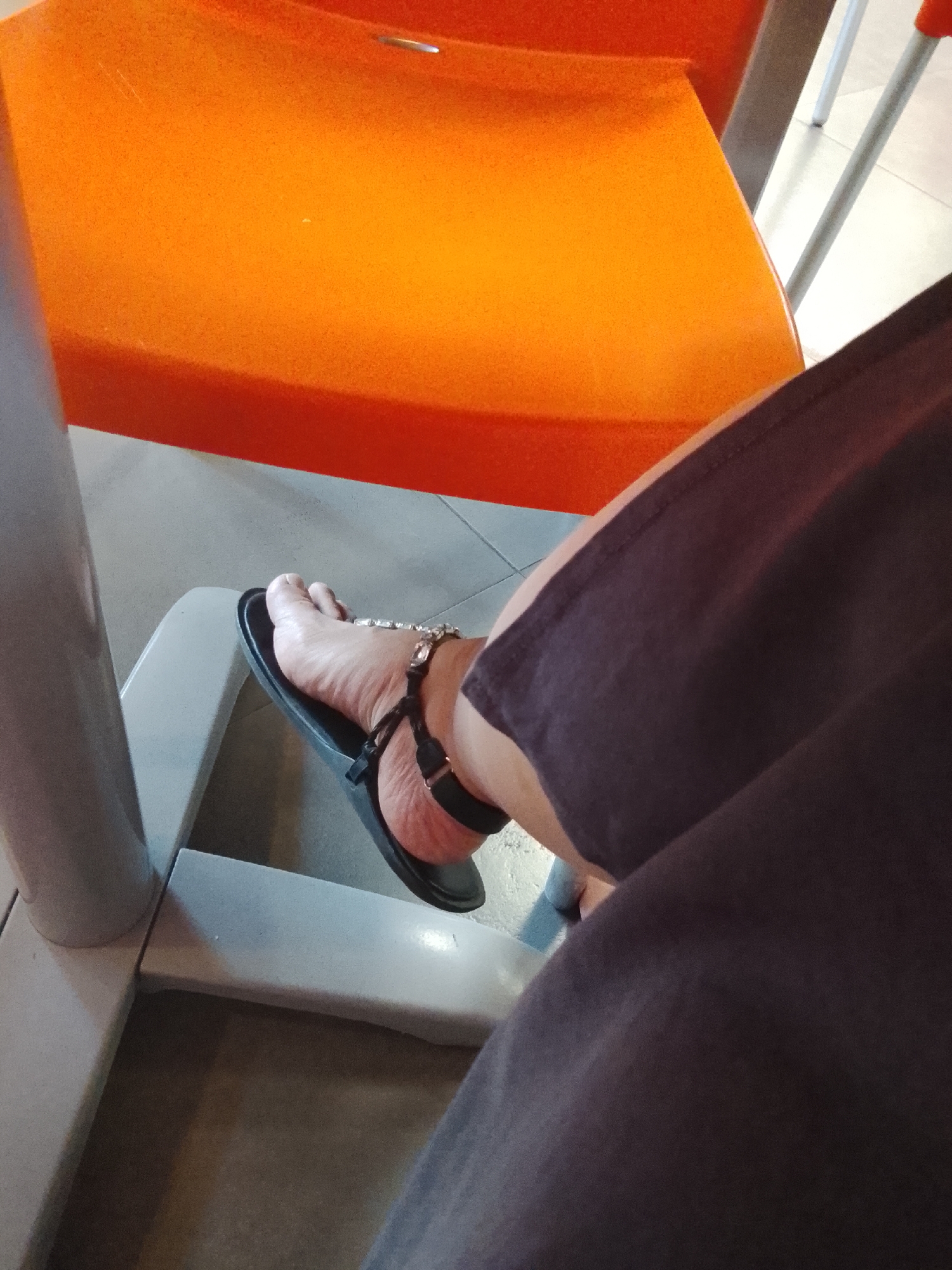 A person's bare foot is shown wearing a black sandal with a rhinestone embellishment. The foot is resting on a light-colored, angular structure, possibly part of a chair or bench. In the background, a bright orange surface, likely a chair seat, is visible. The environment appears to be indoors with a tiled floor. The image does not contain sufficient information to determine the specific location within Almaty, Kazakhstan.