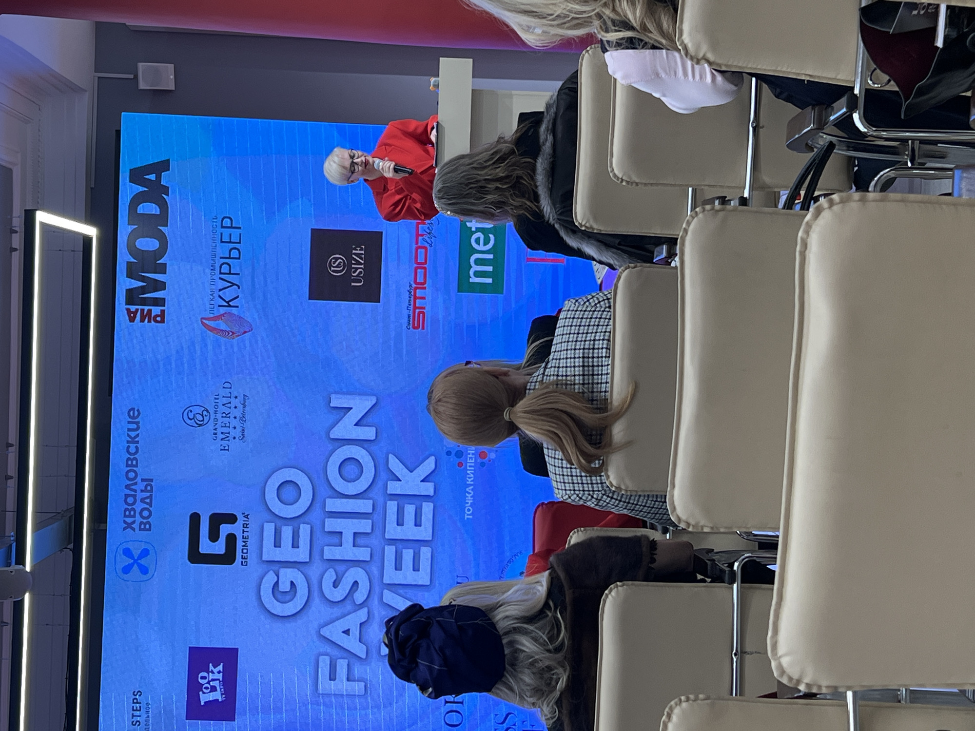 The image shows an indoor event in Saint Petersburg, Russia, likely a fashion-related conference or presentation, judging by the large screen displaying the words "GEO FASHION WEEK" and the logos of several sponsors, including "GRAND-HOTEL EMERALD Saint Petersburg." There are rows of empty beige chairs in the foreground, focusing the view on the attendees and the speaker on stage. The speaker is a woman with short blonde hair and glasses, dressed in a red outfit, holding a microphone. The audience is partially visible, mainly showing the backs of their heads and clothing. The overall atmosphere seems formal and professional.