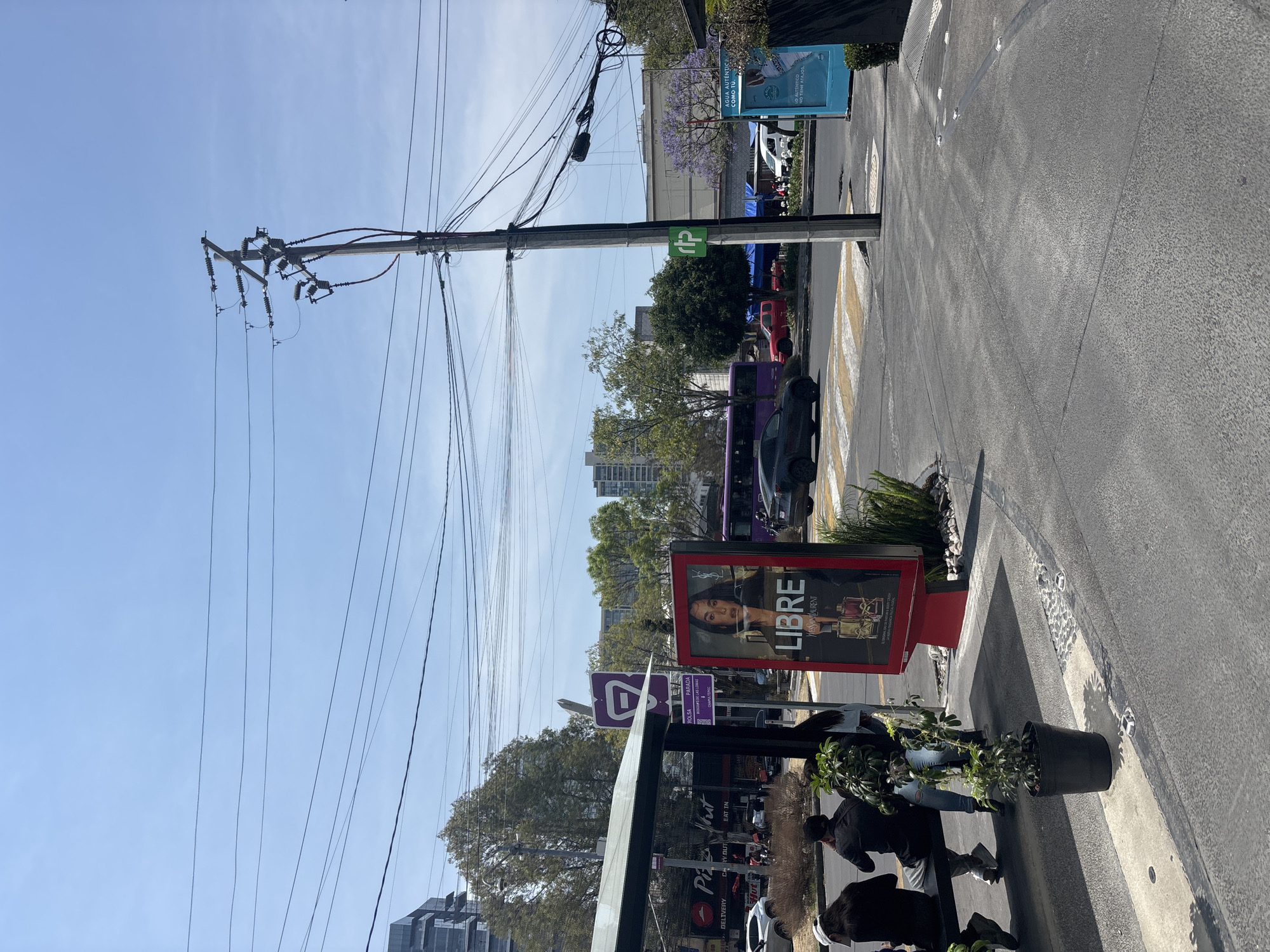 The image depicts a street scene in Mexico City, Mexico. A utility pole laden with electrical wires stands prominently in the foreground, contrasting with the clear blue sky. Adjacent to the pole, a bus stop shelter is visible, adorned with advertisements for "Libre YSL," a fragrance, and "Pizza Hut." A purple and white "rtp" bus stop sign is near the shelter. Pedestrians, partially obscured, can be seen within and around the bus stop. A crosswalk marks the roadway, where a purple bus and other vehicles are parked. Beyond the immediate foreground, a mix of buildings and trees populate the cityscape. Additionally, a blue advertisement banner is partially visible further up the street.
