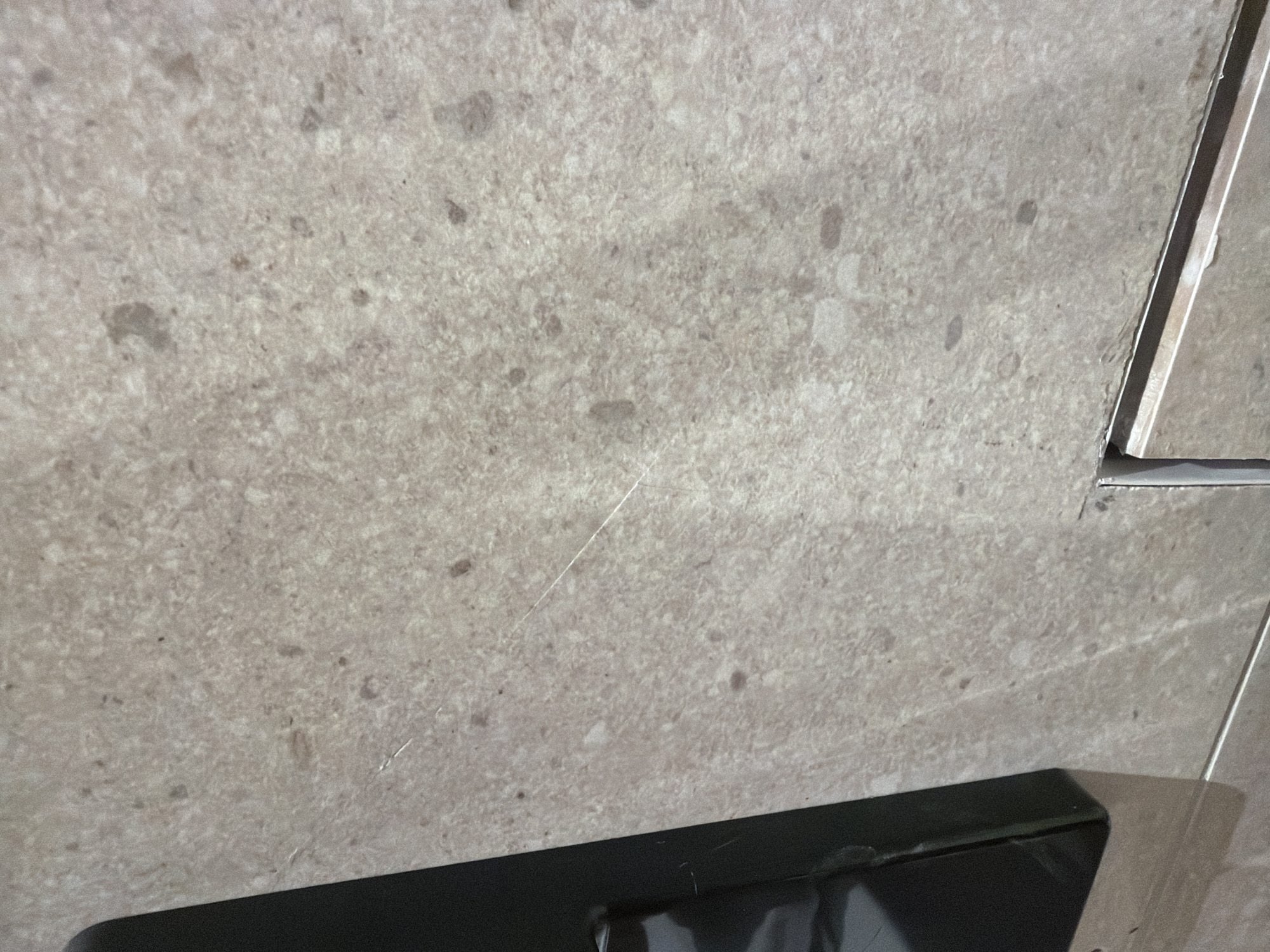 The image shows a close-up view of a textured, light-colored surface, likely tile or stone, with darker speckles. A thin, reflective scratch is visible across the surface. On the left side of the image, a portion of a dark, rectangular object, possibly a piece of electronic equipment or furniture, is partially visible. In the lower portion of the image, another tile or surface is visible below the main textured surface, with a thin metal strip separating them. The lighting suggests an indoor environment. The scene is not identifiable as being in Jerusalem, Israel, or any specific city.