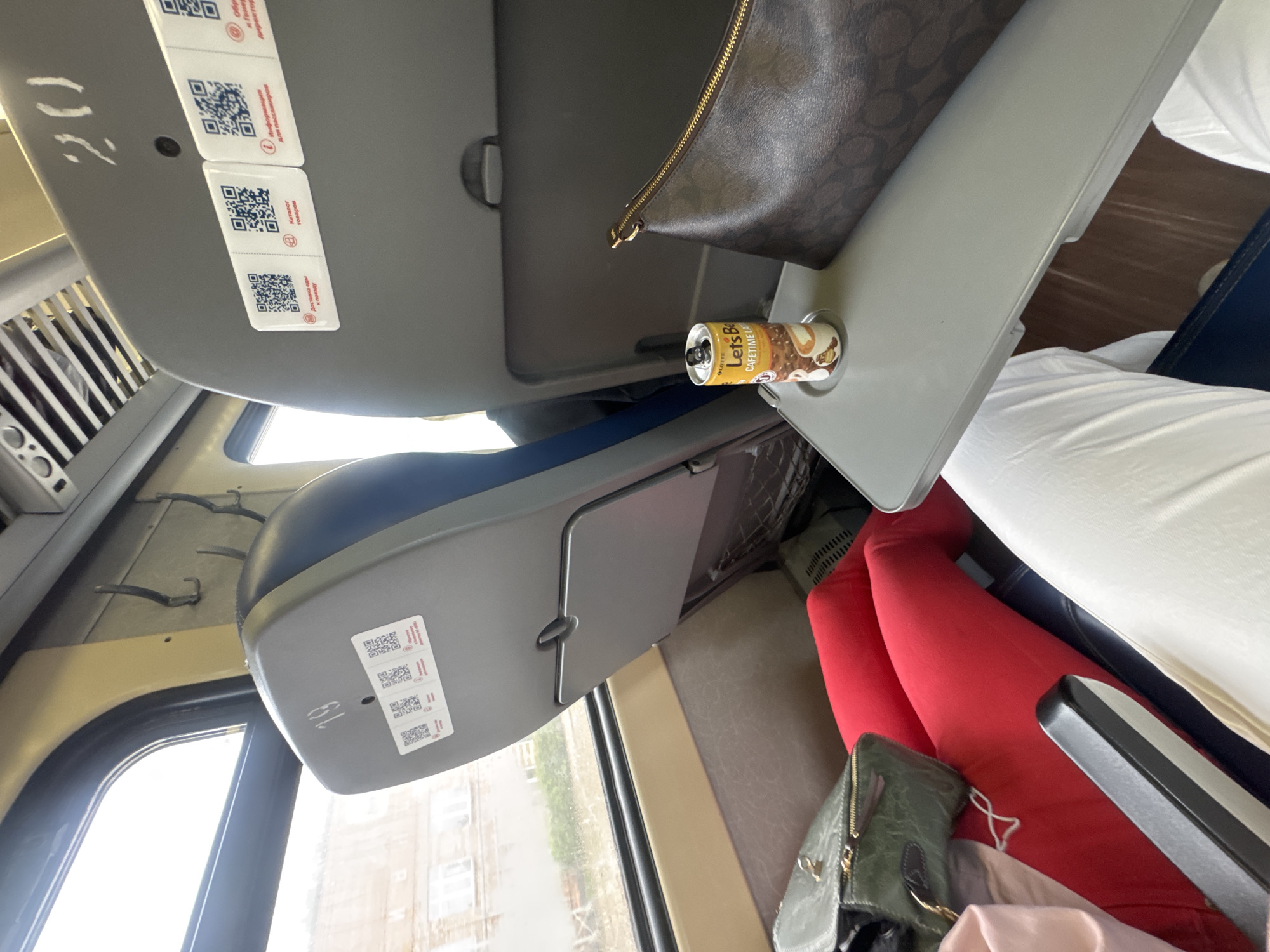 The image depicts the interior of a train carriage, likely a passenger train, with a focus on a tray table. The tray table is deployed and holds a can of "Let's Be CAFETIME LATTE" beverage and a brown patterned clutch purse. In the background, to the left, is a window showing a blurred outdoor scene with buildings and vegetation, suggesting it is daytime. The seats are visible, one with the number "19" and the other with "20" painted on it, along with several QR codes. A person's legs, clad in red leggings and white trousers, are visible, indicating the presence of at least two passengers. The overall environment is functional and utilitarian, typical of public transportation. The specific location within Ussurijsk, Russia cannot be determined from the visual information provided.