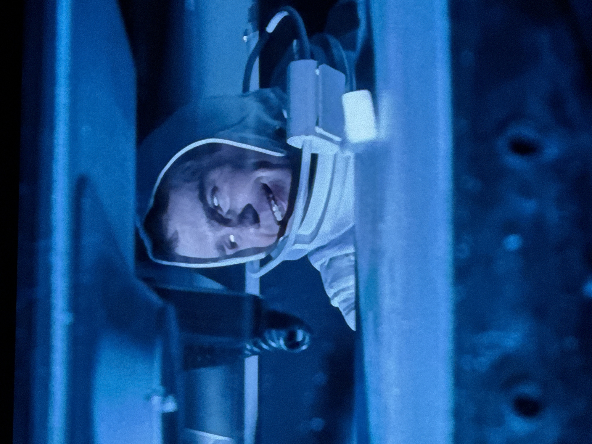 The image displays a man in an enclosed, dimly lit, blue-toned interior, captured from the chest up. The man, appearing to be middle-aged, has short dark hair and is wearing a white or light-colored spacesuit or specialized protective attire, which includes a distinct structured collar around his neck and a hood-like element framing his head. A mechanism consisting of cylindrical and flat components is attached to the right side of his suit's collar. He is looking upwards and slightly to the right with a wide, open-mouthed grin, revealing his teeth, and his eyes are also directed towards the upper right.
Above the man, dark, metallic-appearing structures are visible, including a pipe or beam on the left with a coiled spring-like component attached. A broad, flat structure spans the upper frame, obscuring the ceiling. The foreground shows a dark, textured surface, possibly a console or interior panel. The overall lighting is predominantly blue, creating a cool and artificial ambiance.
There are no identifiable visual cues within the image to determine a specific location in the city of Riga, Latvia, or any other geographic location. The scene appears to be an interior, possibly from a set or vehicle.
