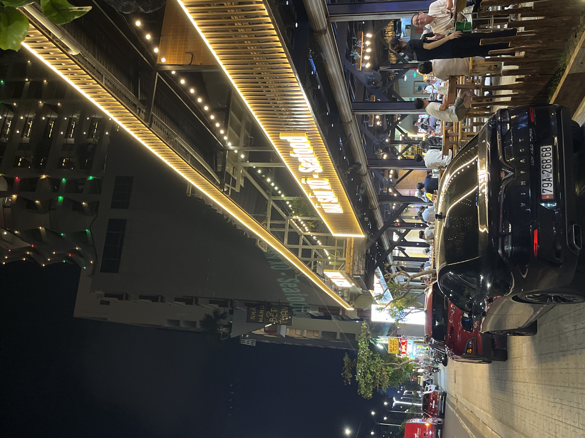 Here is a forensic description of the image:
The image depicts a nighttime scene in Bac Nha Trang Ward, Vietnam, where a restaurant named "Wisnu XO Seafood" is prominently featured. The restaurant is illuminated by a row of horizontal lights and string lights, which illuminate its sign and outdoor dining area. The outdoor seating section is populated by people sitting at tables. A black Vinfast LUX A20 Turbo car with the license plate "79A 268.68" is parked near the dining area. Other vehicles are parked along the street, including a red car directly behind the Vinfast. The street is paved with stone or concrete tiles. The environment includes trees and other buildings, with streetlights and more building lights visible in the distance.