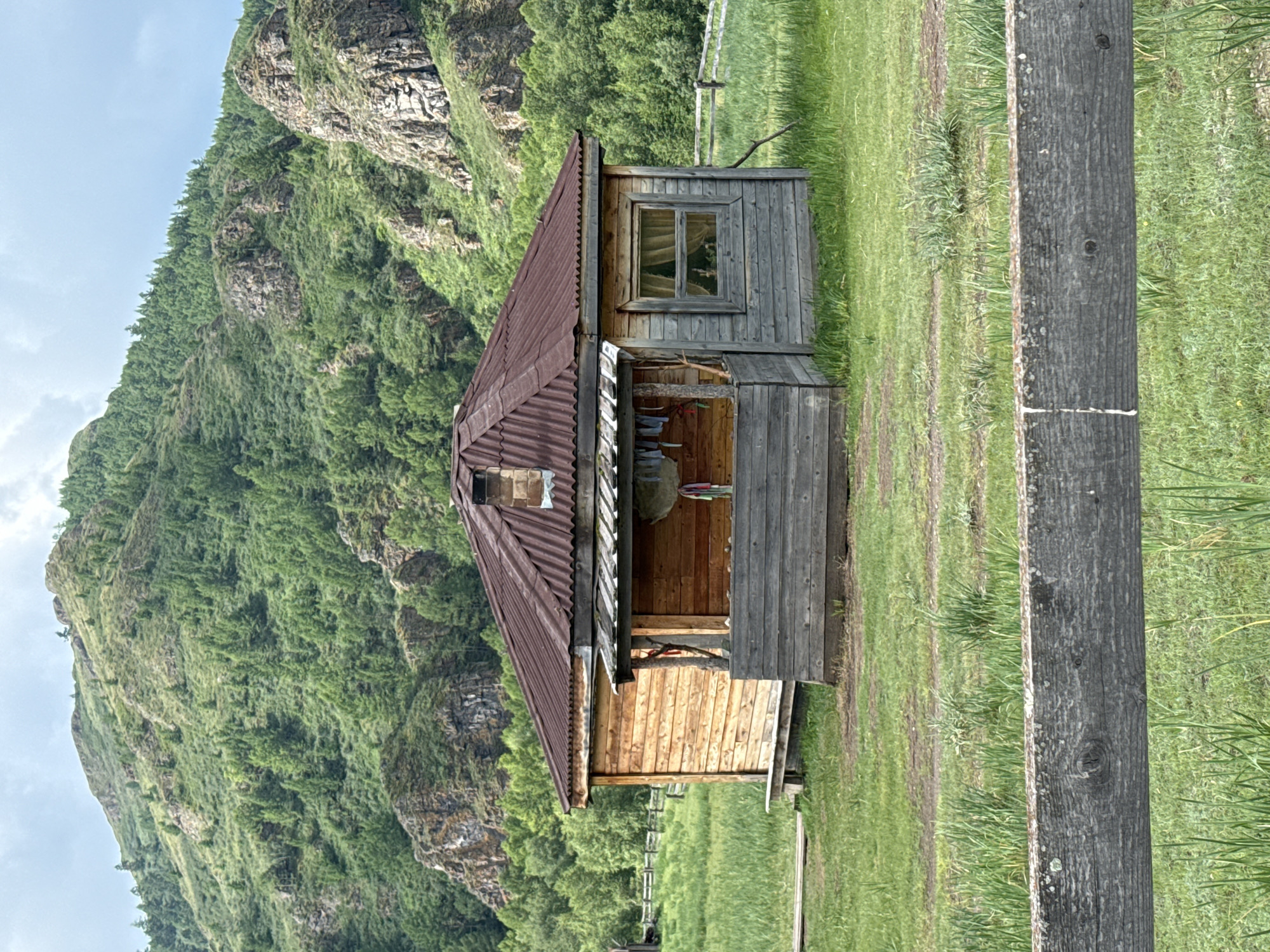 The image depicts a rustic wooden cabin situated in a grassy field with a lush, green mountainous backdrop. The cabin has a corrugated metal roof and a porch area. Several items are hanging on the porch, possibly laundry. A window with curtains is visible on the right side of the cabin. The foreground shows a weathered wooden fence. The environment suggests a rural or remote setting. The location is identified as Kazanovka, Russia.